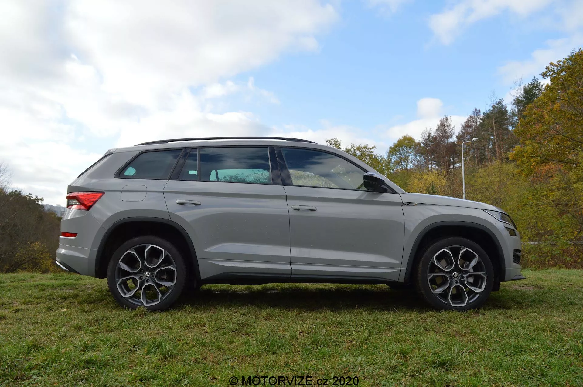 Celkový bočný pohľad na strieborný SUV Skoda Kodiaq 2017 zaparkovaný na tráve s pozadím stromov a zamračenej oblohy. Dizajn vozidla sa vyznačuje výraznými líniami, veľkými zliatinovými kolesami a charakteristickými viaclúčovými diskami. Zrkadlá, ktoré majú integrované smerové svetlá, sú namontované tesne pod líniou okien. Okna sú mierne tónované a línia strechy sa jemne skláňa k zadnej časti, čo naznačuje športový, kupéový profil. SUV je tiež vybavené strešným zábradlím.