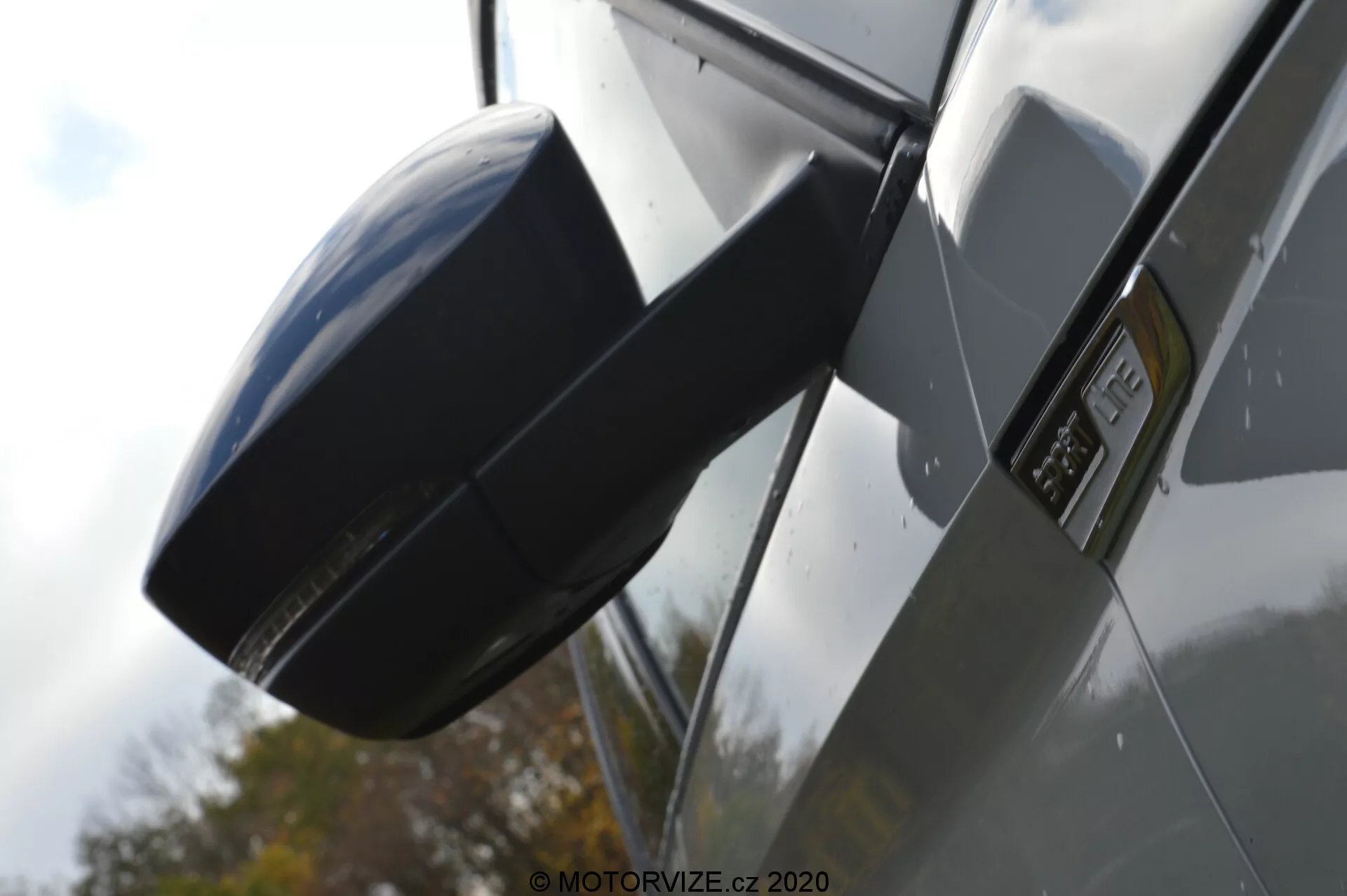 Detailný pohľad na bočné spätné zrkadlo a oblasť dverí vodiča vozidla Škoda Kodiaq 2017 s dôrazom na moderný aerodynamický dizajn s lesklými lakovanými povrchmi a možnou čiernou plastovou alebo uhlíkovou textúrou, zdôrazňujúci zrkadlo a emblém na dverách. Pozadie je rozmazané, čím sa zvyšuje detail zrkadla pod zatiahnutým osvetlením.