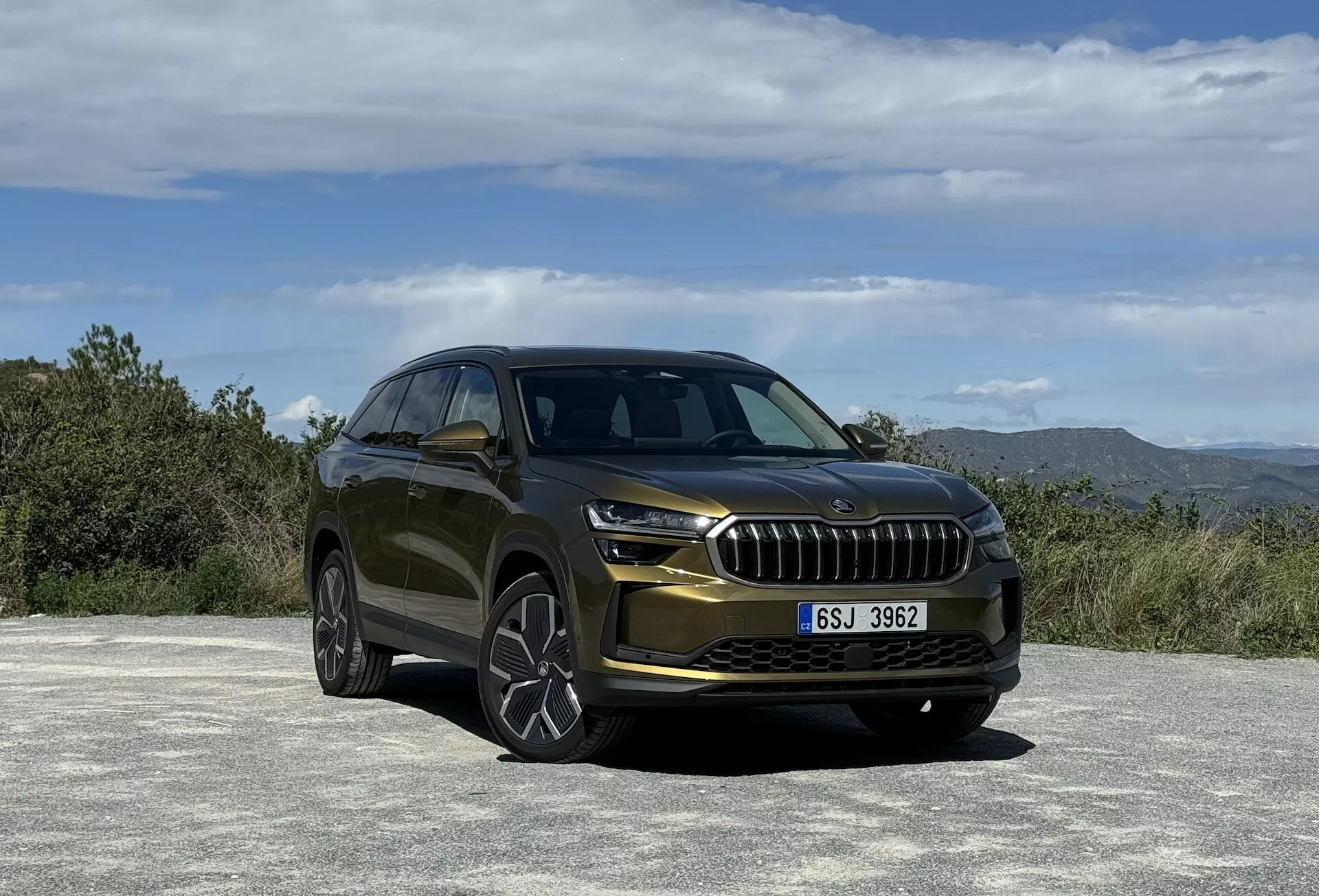 SUV Skoda Kodiaq 2024 v trojštvrtečnom pohľade spredu zdôrazňujúci jeho luxusnú mriežku, štýlové LED svetlomety, športové puzdrá hmlových svetiel a vzduchové vstupy, umiestnené proti prírodnej krajine s metalickým lakom a viacerými hovorenými zliatinovými kolesami.