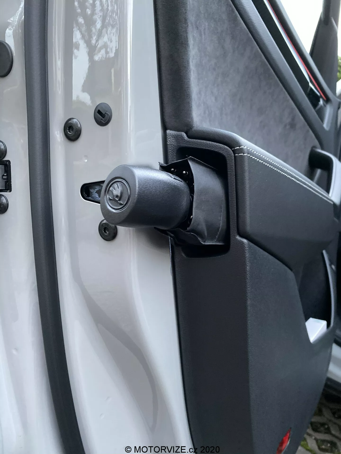 Detailný pohľad na robustný mechanizmus závesu dverí a okolitú oblasť na modeli Škoda Kodiaq 2017, s viditeľnou pružinovou súčasťou pre náročné použitie. Biele vozidlo kontrastuje s čiernymi komponentmi závesu vrátane tradičného zámkového mechanizmu s dierkou na kľúč nad závesom, upevneného šesťhrannými skrutkami pre odolnosť. Textúrovaný plast vnútornej časti dverí a zabezpečené montážne body zvyšujú dôraz na úžitkový dizajn. Rozmazané pozadie zvýrazňuje záves a zámkový mechanizmus.
