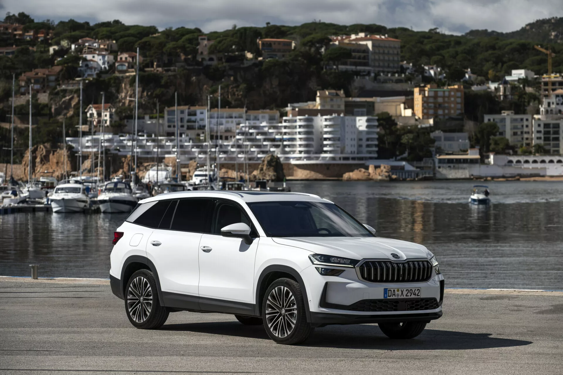 Crossover SUV Škoda Kodiaq 2024 zaparkovaný na nábreží pri mori, zobrazujúci celý vůz vrátane moderných prvkov, ako sú LED svetlomety, veľká mriežka, kolesá z ľahkej zliatiny s viacerými lúčmi, s malebným pozadím lodí a mestského panorámy