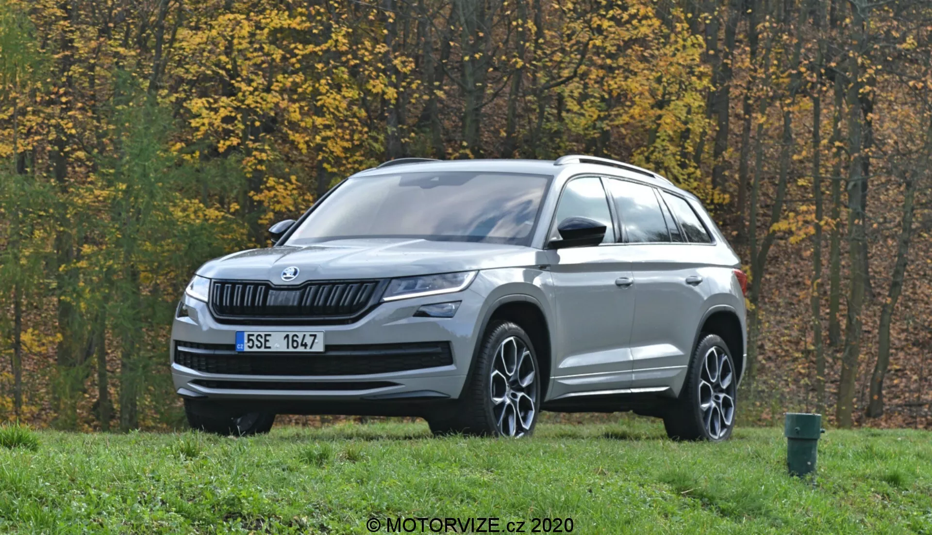 Obrázok zobrazuje SUV Škoda Kodiaq 2017 zaparkované na trávnatej ploche s jesennými stromami v pozadí. Vozidlo je nafotené z trojštvrťového predného pohľadu a má horizontálnu lamelovú mriežku s logom Škoda v strede. Elegantné LED svetlomety sa tiahnu do strán a pod nimi sa nachádzajú hmlovky. Kapota má jemné záhyby a spodná časť predného nárazníka je ozdobená strieborným prvkom. Profil vozidla zahŕňa viacramenné zliatinové kolesá, zrkadlá s integrovanými smerovkami, strešné nosiče a tvarované línie karosérie. Snímače alebo kamery na spodnej časti nárazníka naznačujú pokročilé asistenčné systémy vodiča (ADAS). Venkovné prostredie zahŕňa asfaltovú cestu a zelený odpadkový kôš na pravom okraji záberu.