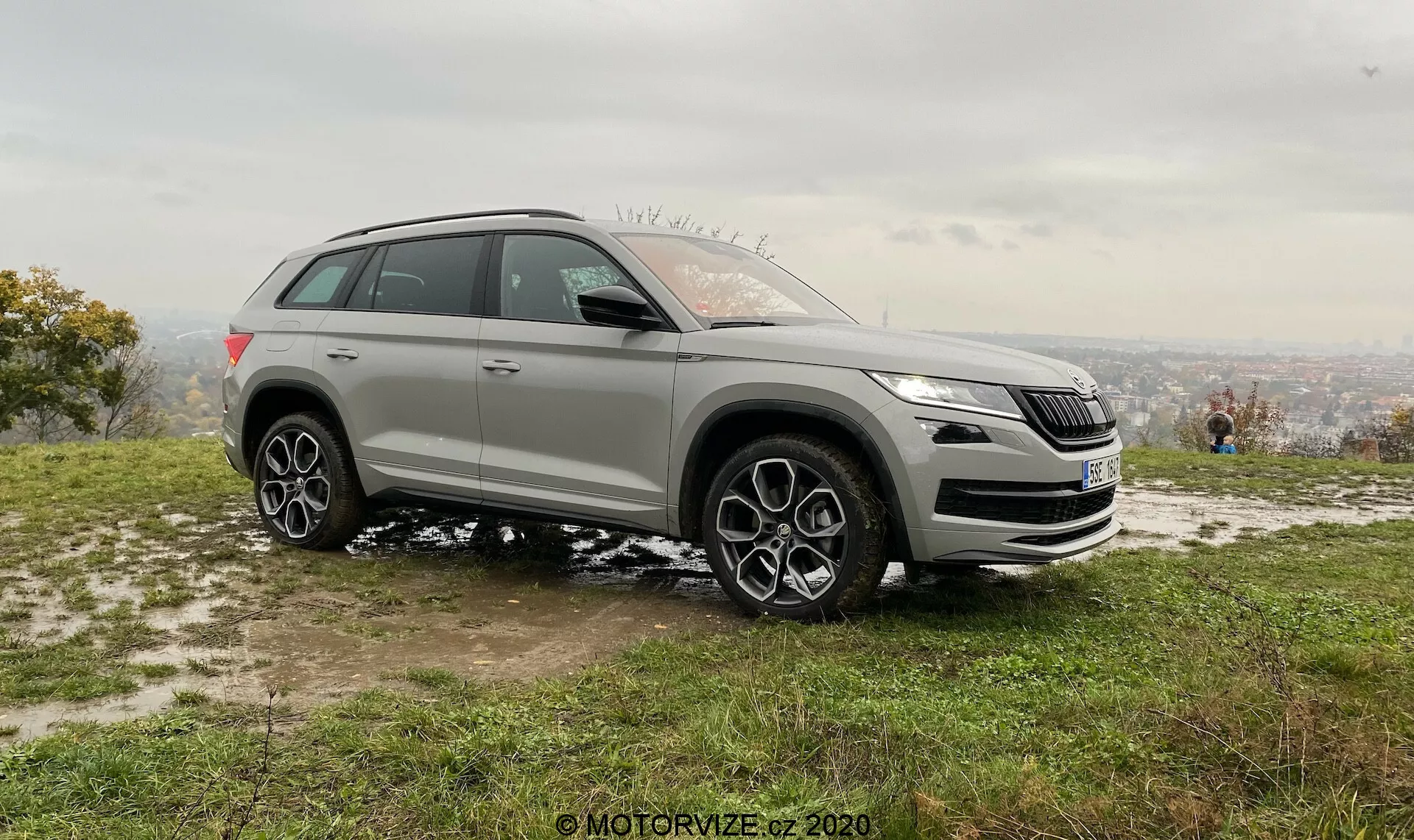 Bočný pohľad na moderné SUV nasnímané z šikmého uhla, zobrazujúci ako predné, tak bočné dizajnové prvky, zasadený do prírodného pozadia z výškového bodu. Obrázok zvýrazňuje prednú masku, svetlomety, hmlové svetlá a charakteristické logo vozidla. Zobrazuje tiež vysokú svetlú výšku, veľké kolesá z ľahkých zliatin s viacerými lúčmi, výrazné oblúky kolies, vysokú ramenovú líniu, bočné zrkadlá, strešné nosiče a tónované okná. Sportový dizajn a potenciálne pokročilé technológie vozidla sú zvýraznené v tomto stacionárnom dennom zábery s fľakmi snehu, čo naznačuje chladnejšiu sezónu.