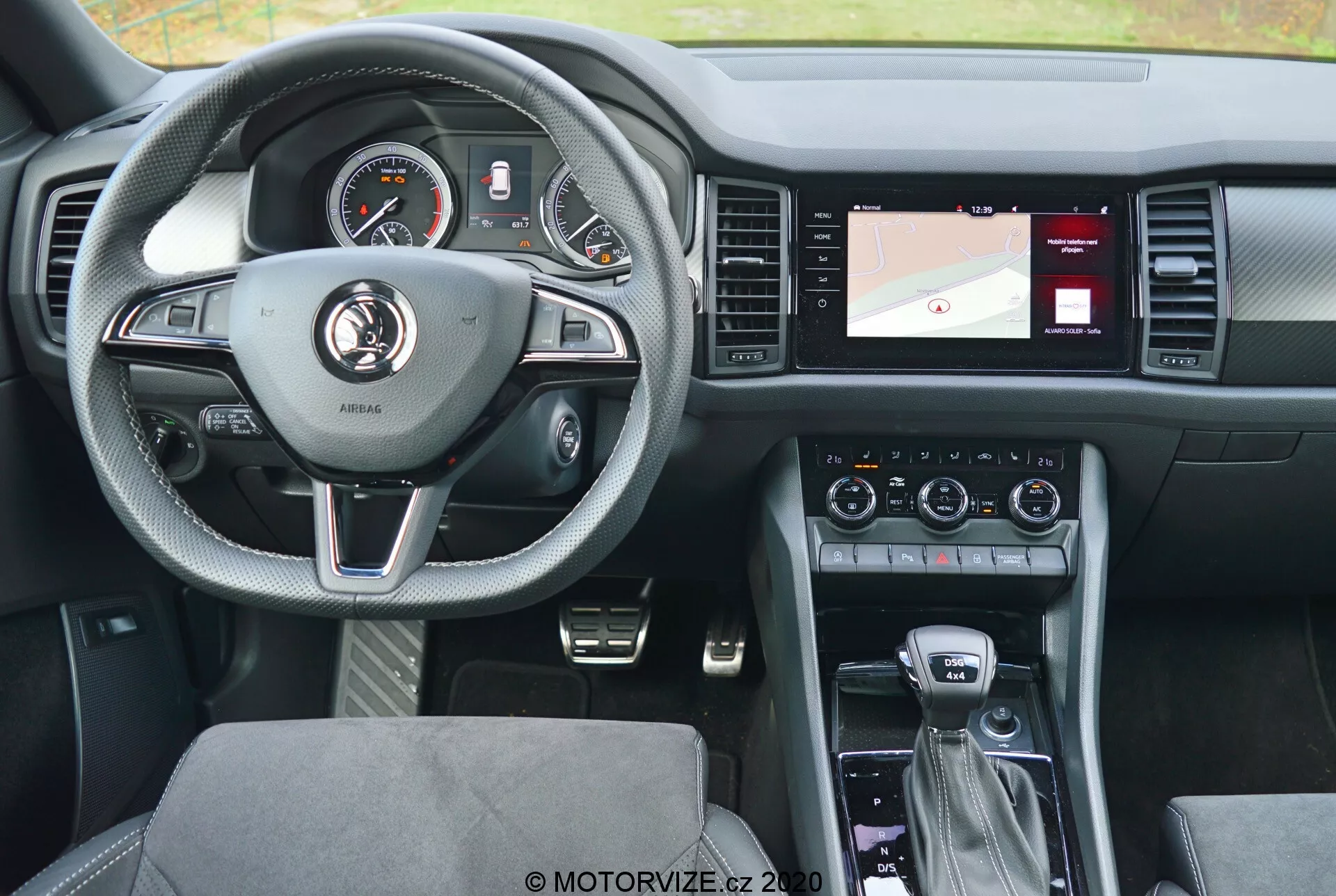 Pohľad na interiér vozidla Škoda Kodiaq 2017 zo zadného sedadla, zvýrazňujúci kožený trojramenný volant so striebornými akcentmi a ovládacími tlačidlami pre tempomat, audio a hands-free komunikáciu. Prístrojový panel za volantom obsahuje ako analógové ciferníky, tak digitálny displej. Stredová konzola vpravo zahŕňa obrazovku s navigáciou, ovládanie klimatizácie a nastavenia audio systému. Radenie naznačuje automatickú prevodovku s ďalšími ovládacími prvkami pre infotainment systém a jazdné režimy. Moderný dizajn smeruje k pohodliu a vyváženosti technológií s čalúnickými sedadlami a dobre osvetleným, užívateľsky prívetivým rozvrhnutím.