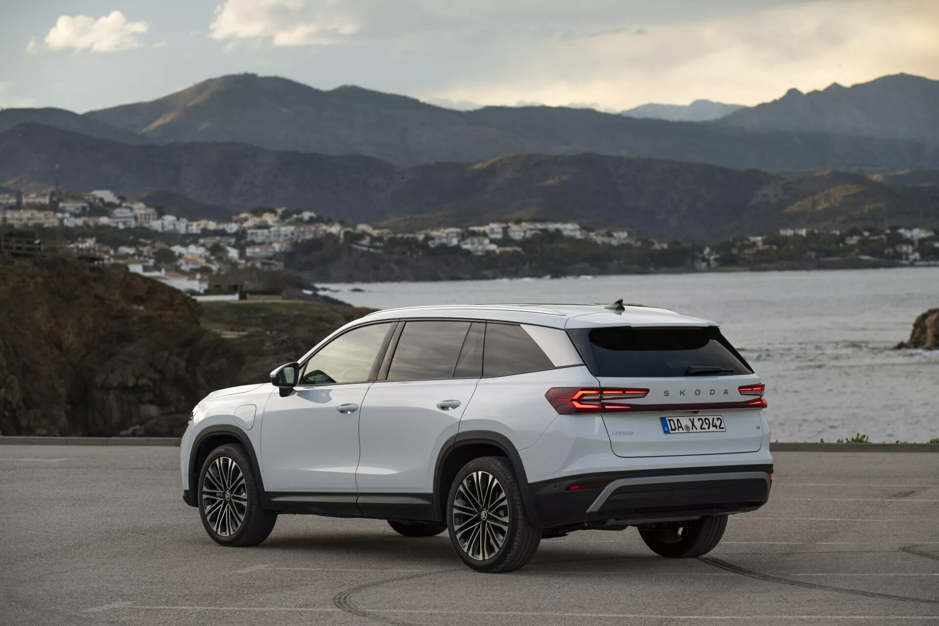 Zadný trojštvrťový pohľad na biely Skoda Kodiaq 2024, predstavujúci bočné a zadné dizajnové línie, viaceré hovorové ľahké disky, LED zadné svetlá a označenie modelu, zaparkovaný vonku na pobrežnej ceste počas zlatej hodiny