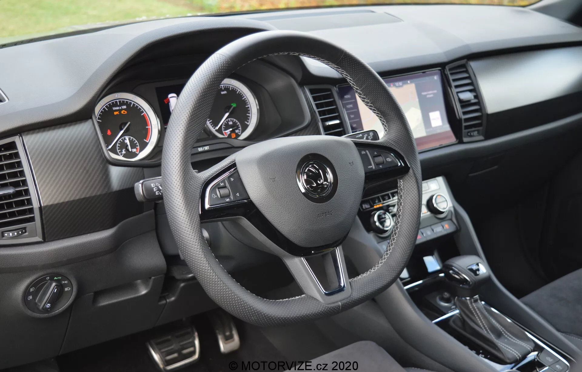 Interiér modelu Škoda Kodiaq 2017 z pohľadu vodiča na prístrojový panel. Trojramenný volant obalený materiálom podobným koži s kontrastným prešívaním má integrované ovládacie prvky pre audio, hands-free komunikáciu a tempomat. Analógové prístroje, s tachometrom vľavo a otáčkomerom vpravo, dopĺňa digitálny displej medzi nimi. Viditeľné sú aj dotyková obrazovka infotainmentu, lemovaná vetracími mriežkami, ovládače klimatizácie a radiaca páka, spolu s textúrovanými materiálmi a brúsenými kovovými akcentmi.