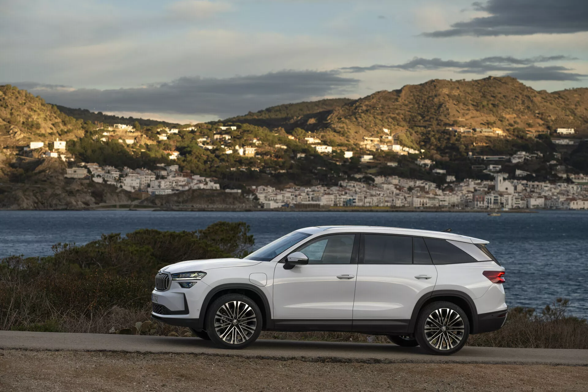 Moderné SUV Skoda Kodiaq 2024 zaparkované na pobrežnej ceste s malebným výhľadom na zátoku a domy na svahu v pozadí, predstavujúce elegantný dizajn, dlhý rázvor, vysokú svetlú výšku, ostre línie karosérie, zapustené kľučky dverí, tónované zadné okná, strešné nosiče, veľké zliatinové kolesá, LED svetlomety, bočné zrkadlá s integrovanými smerovými signálmi a panoramatické strešné okno, všetko na kontrastnom bielom vonkajšom finiše.