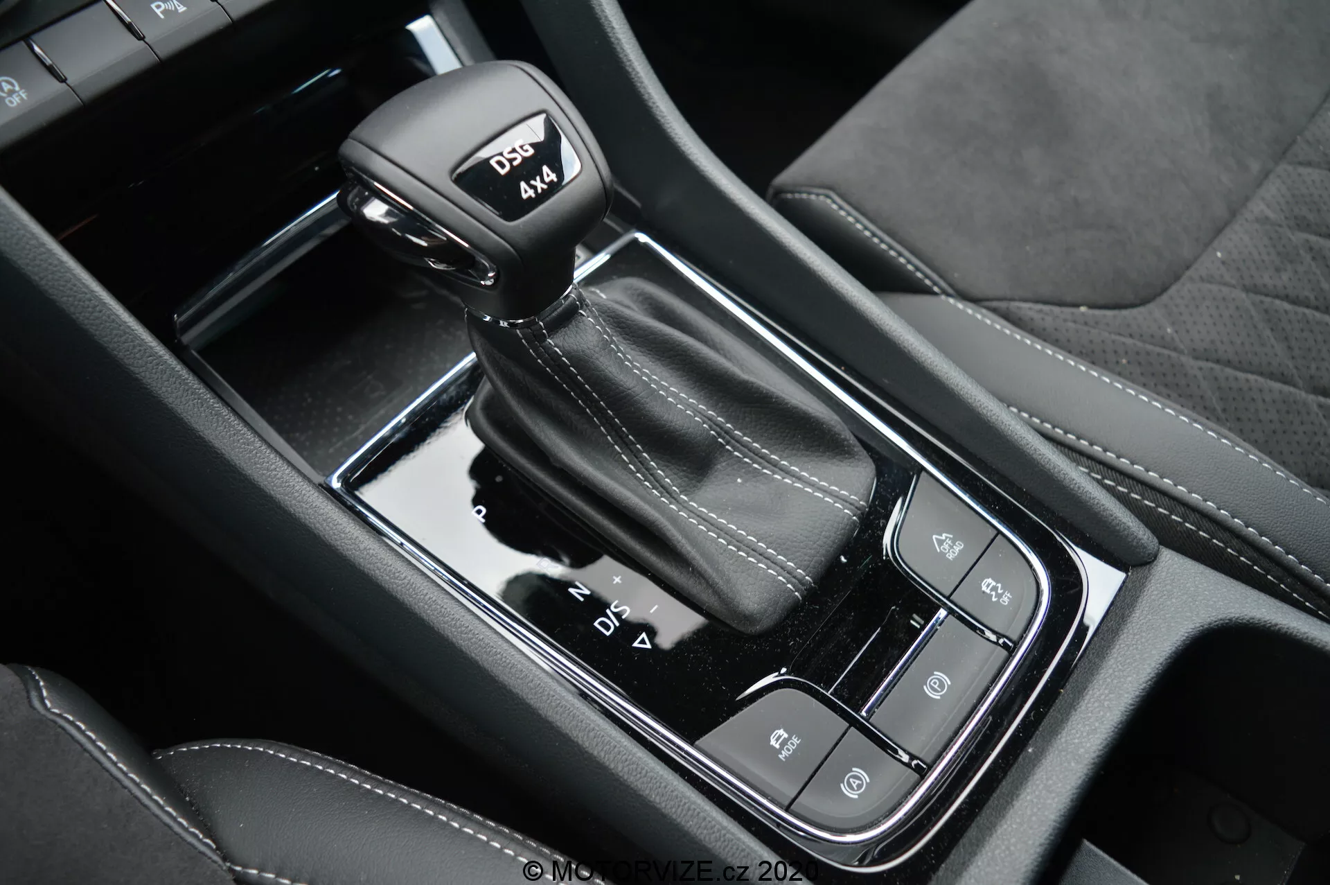 Detailný pohľad na automatickú radiacu páku a lesklú čiernu stredovú konzolu vozidla Skoda Kodiaq 2017, s funkciami jazdných režimov, elektronickej parkovacej brzdy a tlačidlom auto-hold; zdôrazňuje kožené prešívanie na gombíku umožňujúcom športový režim.