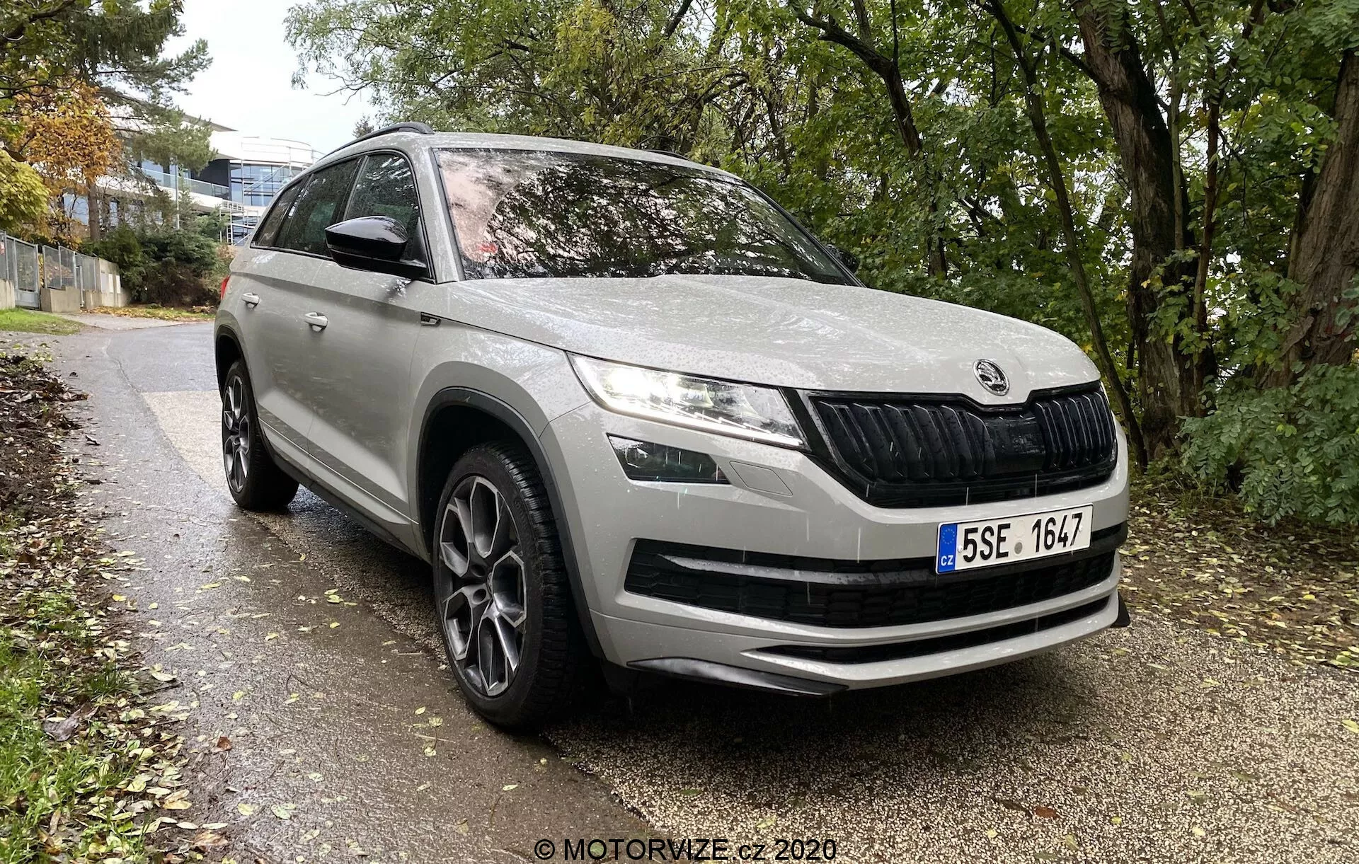 2017 Skoda Kodiaq moderné SUV z európskej automobilovej značky, z predného trojštvrťového uhla ukazuje predný a bočný profil až do stredu zadných dverí, zaparkované pozdĺž chodníka lemovaného trávou a stromami v mestskom alebo prímestskom prostredí. Vlastnosti zahŕňajú charakteristickú prednú mriežku s vertikálnymi lamelami, elegantné LED svetlomety, hmlové svetlá v nárazníku, veľké nasávacie otvory na prednom nárazníku, kontúrové línie na kapote pre aerodynamiku, výrazné oblúky kolies s viaclúčovými zliatinovými kolesami, bočné zrkadlá s integrovanými smerovkami, ochranné lišty na spodnej strane a celkovo čistý, dobre udržiavaný lak.