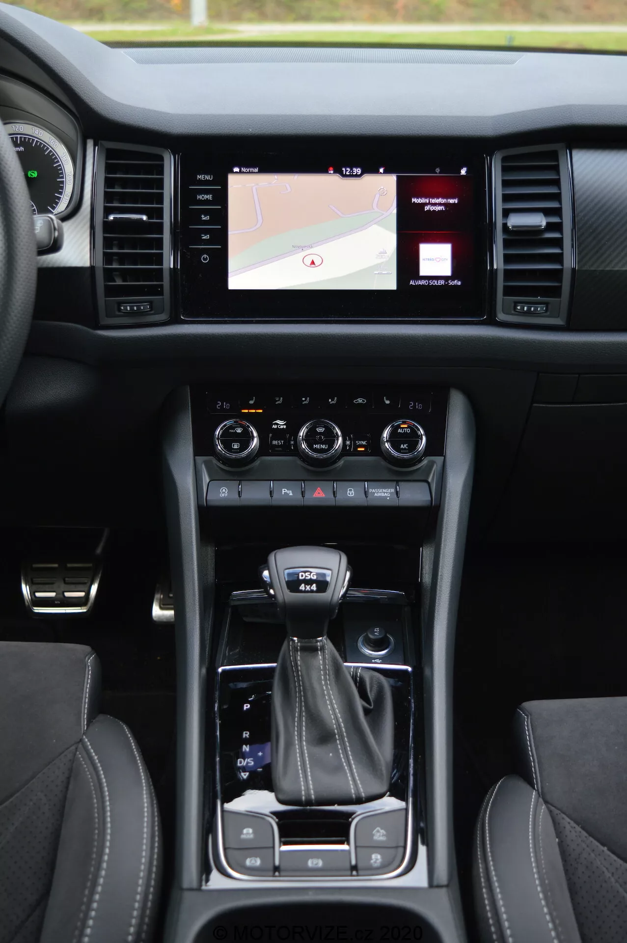 Interiér vozidla Skoda Kodiaq 2017 zobrazujúci centrálnu konzolu, dotykový infotainment systém, zobrazujúci mapu, dvojitý regulátor klimatizácie, radiacu páku, volič jazdných režimov, držiaky na nápoje, kožené sedadlá s kontrastným prešívaním a tlačidlo bezkľúčového zapnutia