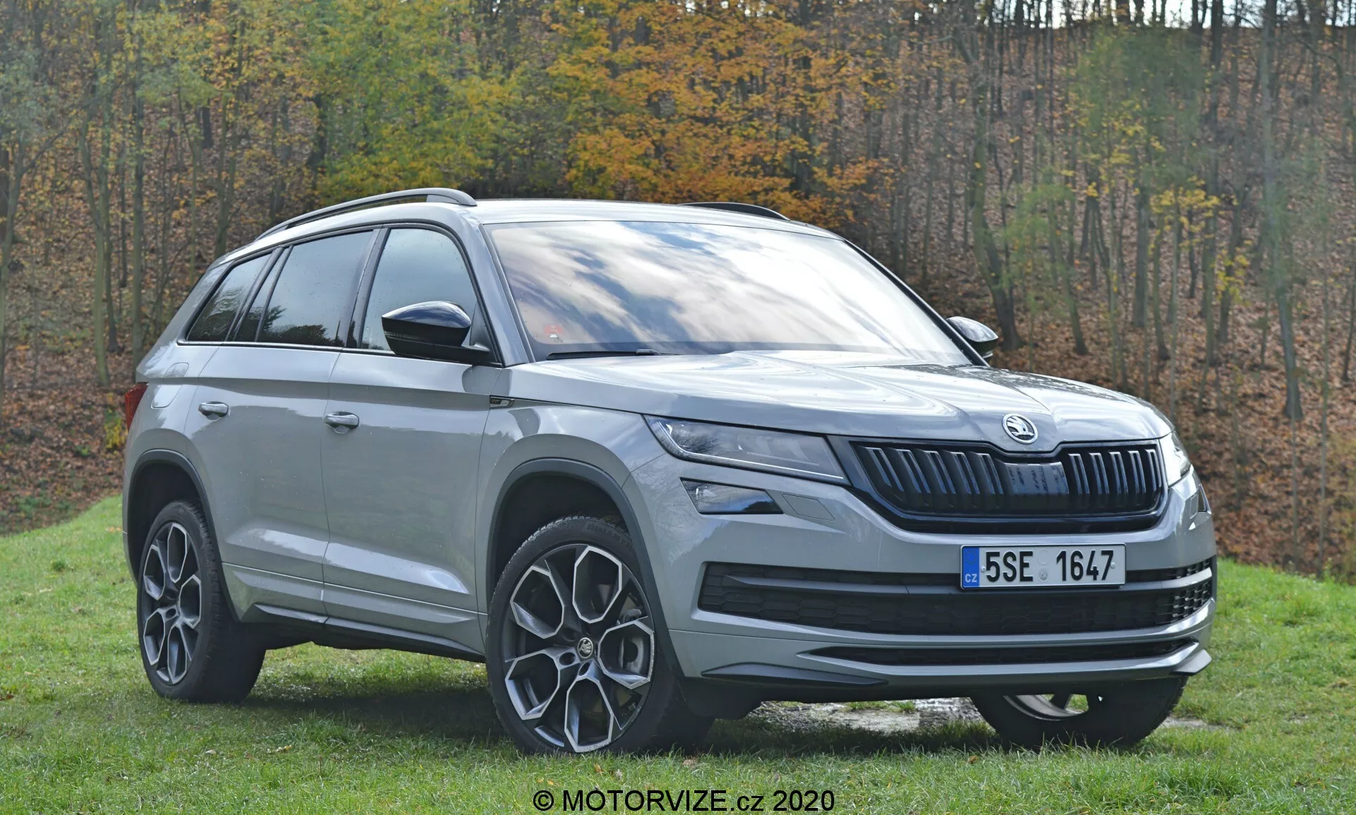 Tento obrázok zobrazuje SUV Škoda Kodiaq z roku 2017 zachytené z trojštvrťového predného uhla, zameraného na ľavú stranu vozidla. Záber zvýrazňuje prednú mriežku s charakteristickým vertikálnym dizajnom Škoda, konfiguráciu svetlometov pravdepodobne s LED technológiou a stredové logo značky. Bočný profil zahŕňa predné dvere vodiča, zadné dvere spolujazdca, strešné nosiče a časti zadnej štvrte. Výrazné zliatinové kolesá s dizajnom lúčov sú tiež výrazne viditeľné. Vonkajšie prostredie s trávnatým pozadím a jesenným lístím zvyšuje estetiku moderného SUV.
