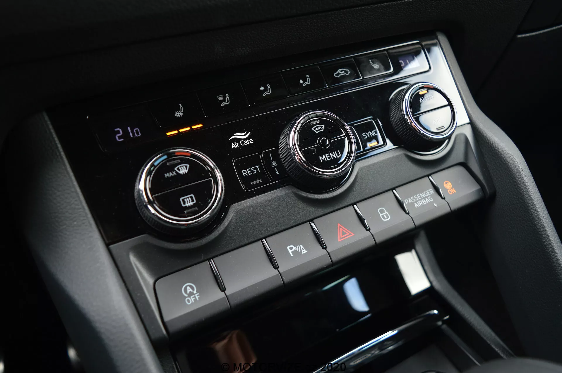 Detailný pohľad na stredovú konzolu vo vozidle Škoda Kodiaq 2017, zvýrazňujúci systém klimatizácie s dvoma veľkými ovládacími gombmi na nastavenie teploty a digitálnym displejom nastaveným na 21 stupňov Celzia. Obrázok tiež obsahuje tlačidlá na klimatizáciu, cirkuláciu vzduchu, odmrazovanie okien a tlačidlo 'MENU'. Nižšie sú prepínače pre parkovacie senzory, deaktiváciu automatického štartu/stop a výstražné svetlá.