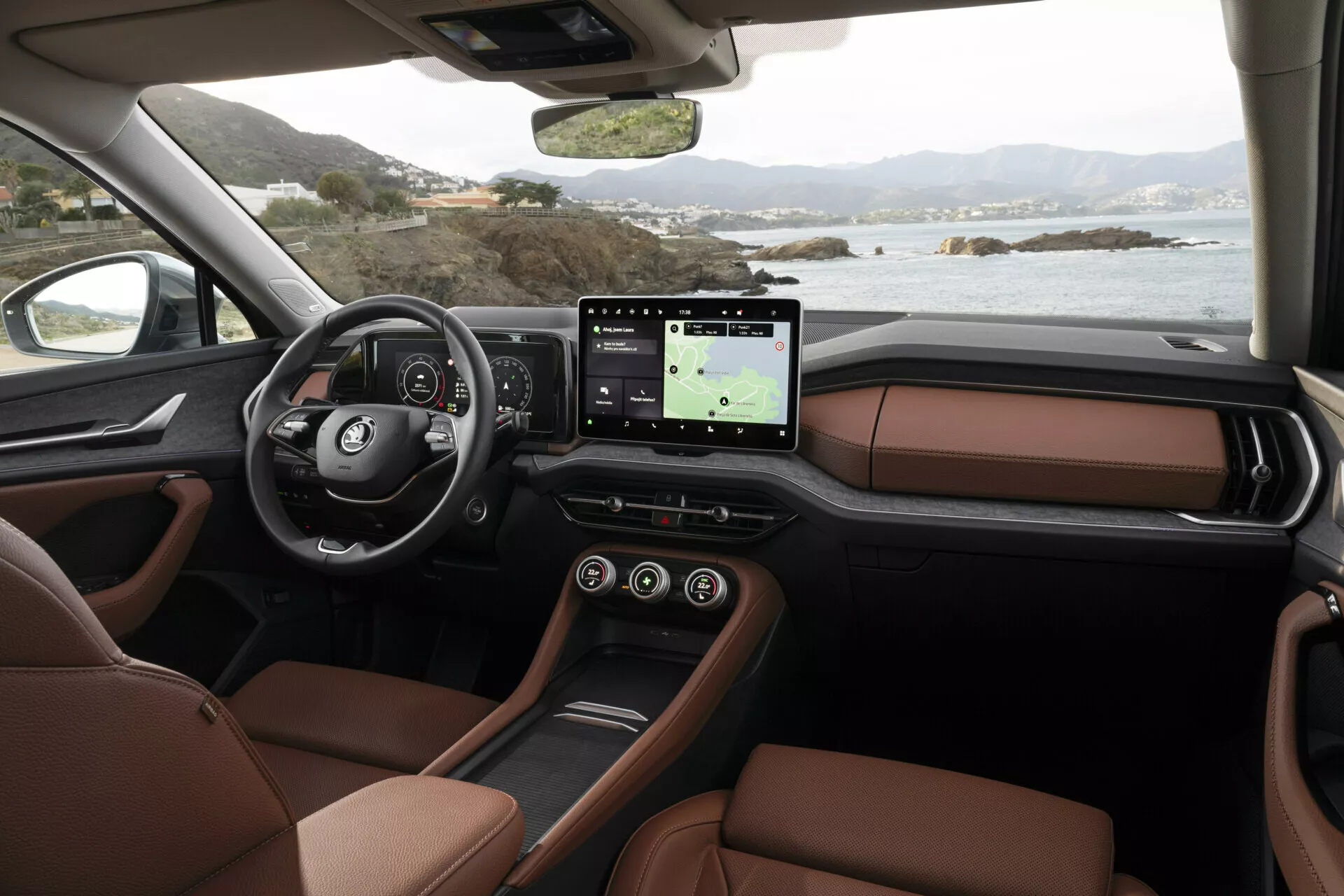 Interiér Skoda Kodiaq 2024 z pohľadu spolujazdca predstavujúci prístrojovú dosku, multifunkčný volant s logom, digitálny prístrojový panel, veľký dotykový infotainment systém s GPS, luxusné hnedé kožené sedadlá a štýlový dizajn stredovej konzoly, so scenériou pokojnej pobrežnej krajiny za oknami