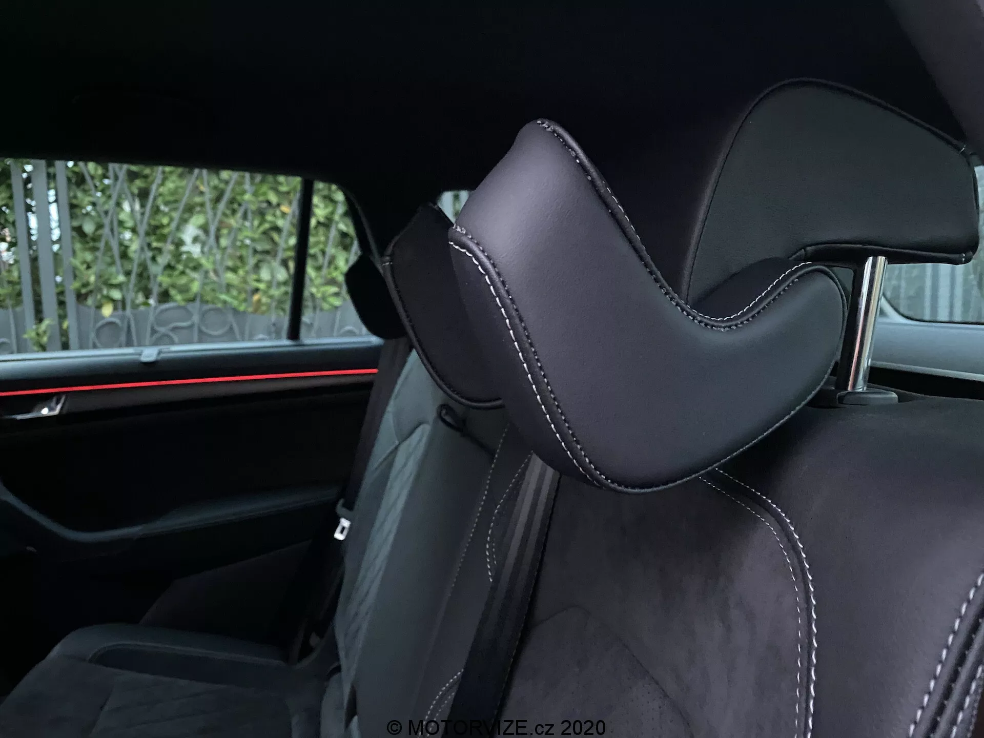 Pohľad na interiér vozidla Skoda Kodiaq 2017 zameraný na sedadlo. Uhol snímky zdôrazňuje časť opierky hlavy, čalúnenú čiernym materiálom, pravdepodobne kožou alebo vysoko kvalitným syntetickým materiálom, s výrazným tvarovaním pre pohodlie. Precízne šitie kontrastnou niťou pridáva luxusné detaily. V pozadí je viditeľné horizontálne červené ambientné osvetlenie, ktoré zdôrazňuje moderné prvky automobilového dizajnu. Viditeľné vybavenie pre zadných pasažierov zahŕňa bezpečnostné pásy a možno aj stredovú opierku, čo naznačuje druhý rad sedadiel.