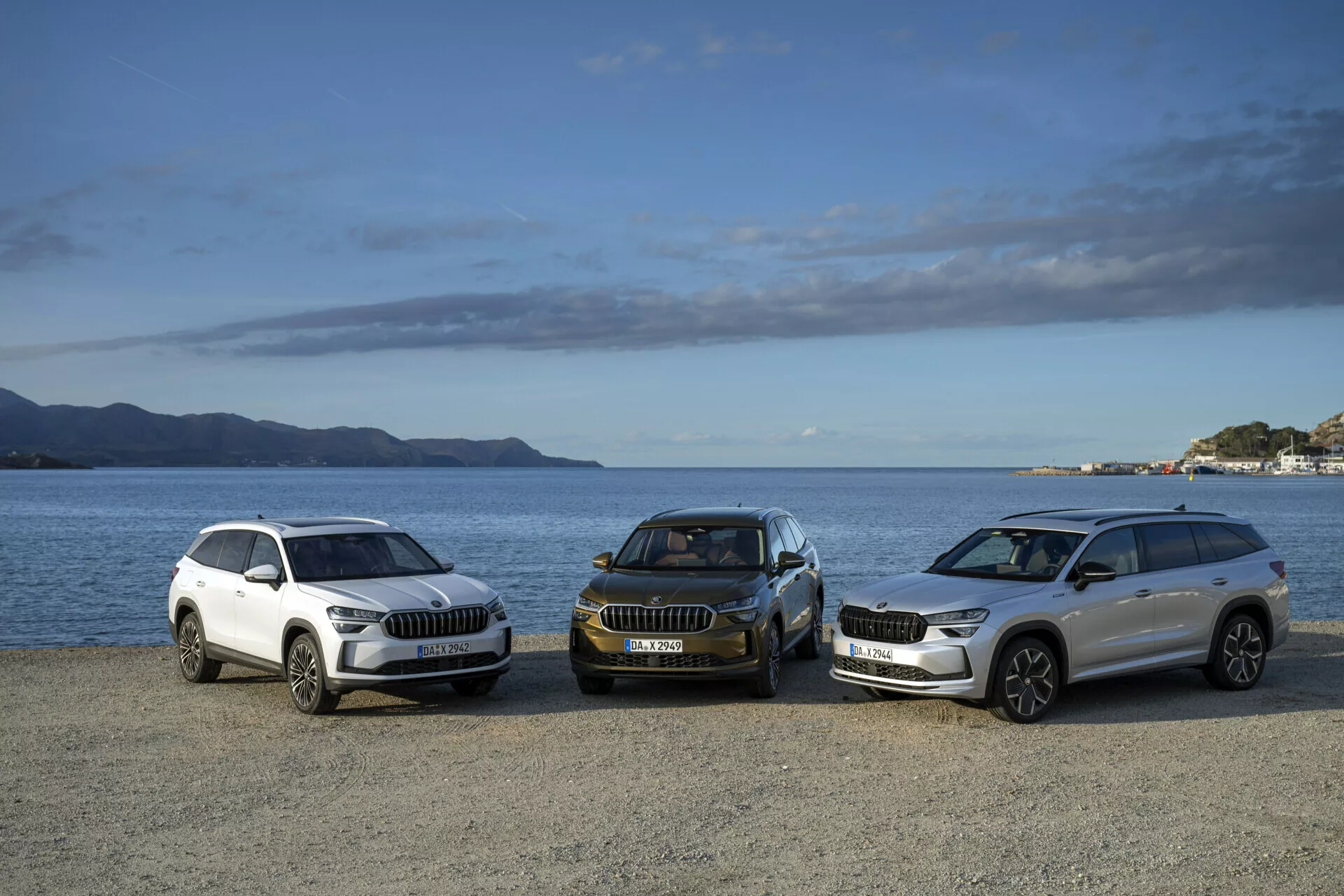 Rada SUV Skoda Kodiaq 2024 v bielych a tmavých farbách v pohľade spredu a zboku, zaparkované na nábreží s horami a čiastočne zamračeným nebom na pozadí