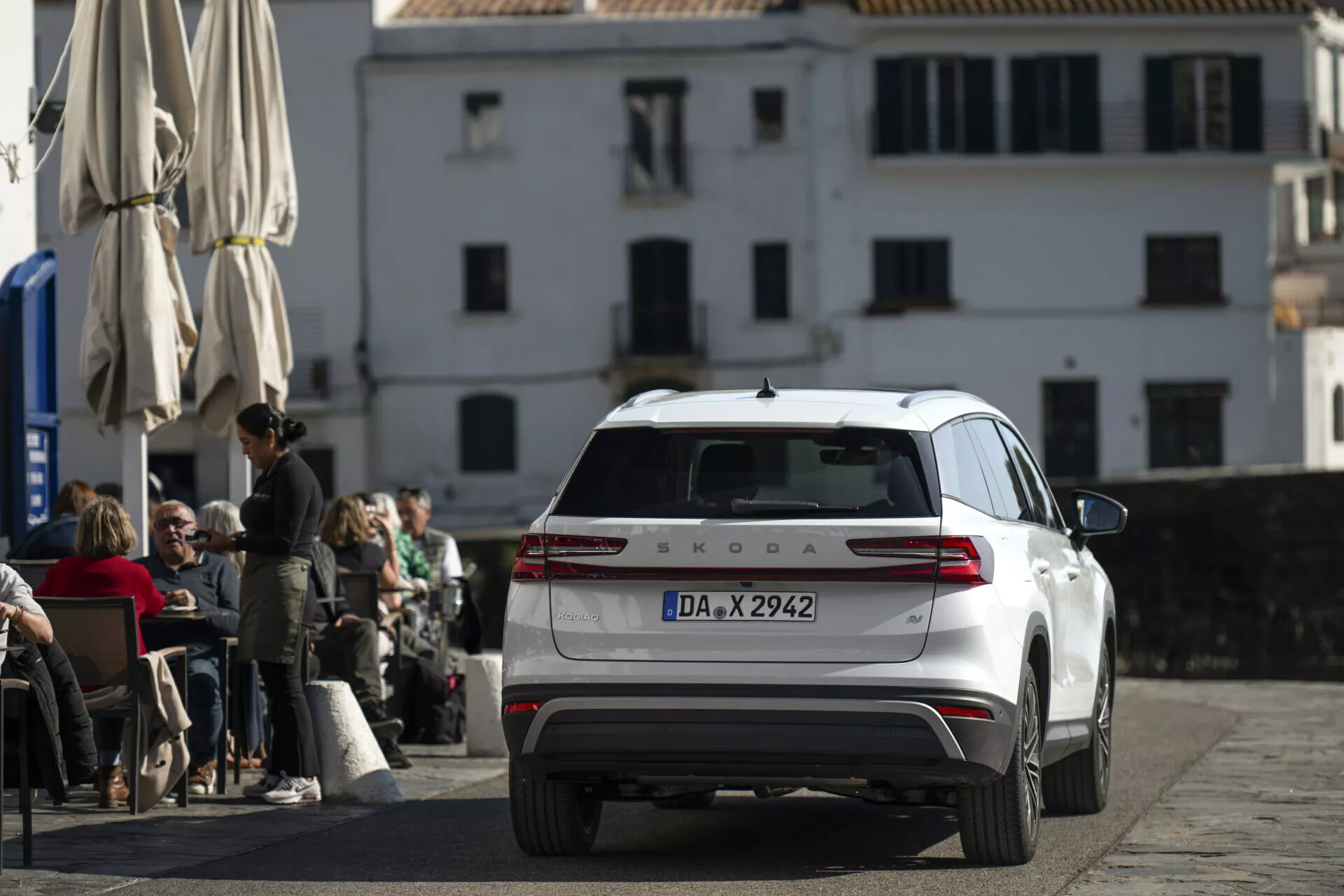 Biely Škoda Kodiaq 2024 fotografovaný z zadného, mierne zvýšeného uhla, zobrazujúci celú zadnú časť, zadné svetlá, veko batožinového priestoru, zadné okno s stieračom a strechu s anténou a nosičmi, zaparkovaný na dlaždenej ulici pred kaviarňou s klasickou európskou architektúrou na pozadí.