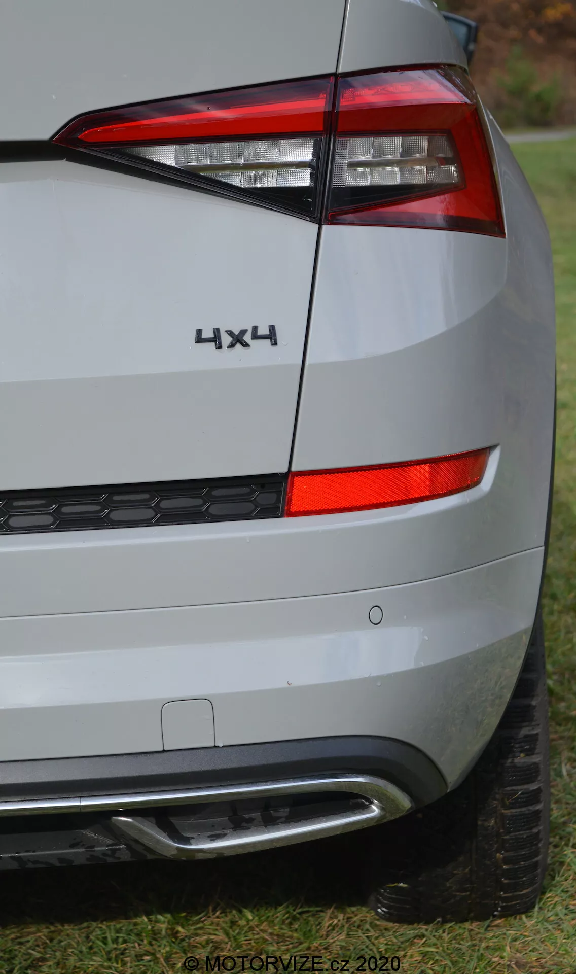 Detailný pohľad na zadný roh vozidla Skoda Kodiaq 2017, zameraný na zadné osvetlenie, označenie a dizajn nárazníka. Uhol záberu zdôrazňuje pravú stranu zadnej časti vozidla, ktorá zobrazuje horizontálne usporiadané, ostré, hranaté zadné svetlo obklopujúce bok. Pod zadným svetlom je vidieť zadný panel a nárazník, vo farbe striebornej alebo svetlo šedej, ktorý odráža tlmené prostredie. Nárazník obsahuje čiernu plastovú spodnú časť s včelím vzorom a strieborným alebo chrómovým prvkom, pravdepodobne súvisiaci s asistenčným systémom parkovania alebo dizajnom. Značka '4X4' nad zadným svetlom označuje pohon všetkých štyroch kolies. Vysokorozlíšený obrázok odhaľuje detailné textúry povrchu, čiastočne viditeľný dezén pneumatiky na dne, umiestnený na trávniku. Vodotlač '© MOTORVIZE.cz 2020' naznačuje, že obrázok je určený pre českú automobilovú publikáciu.