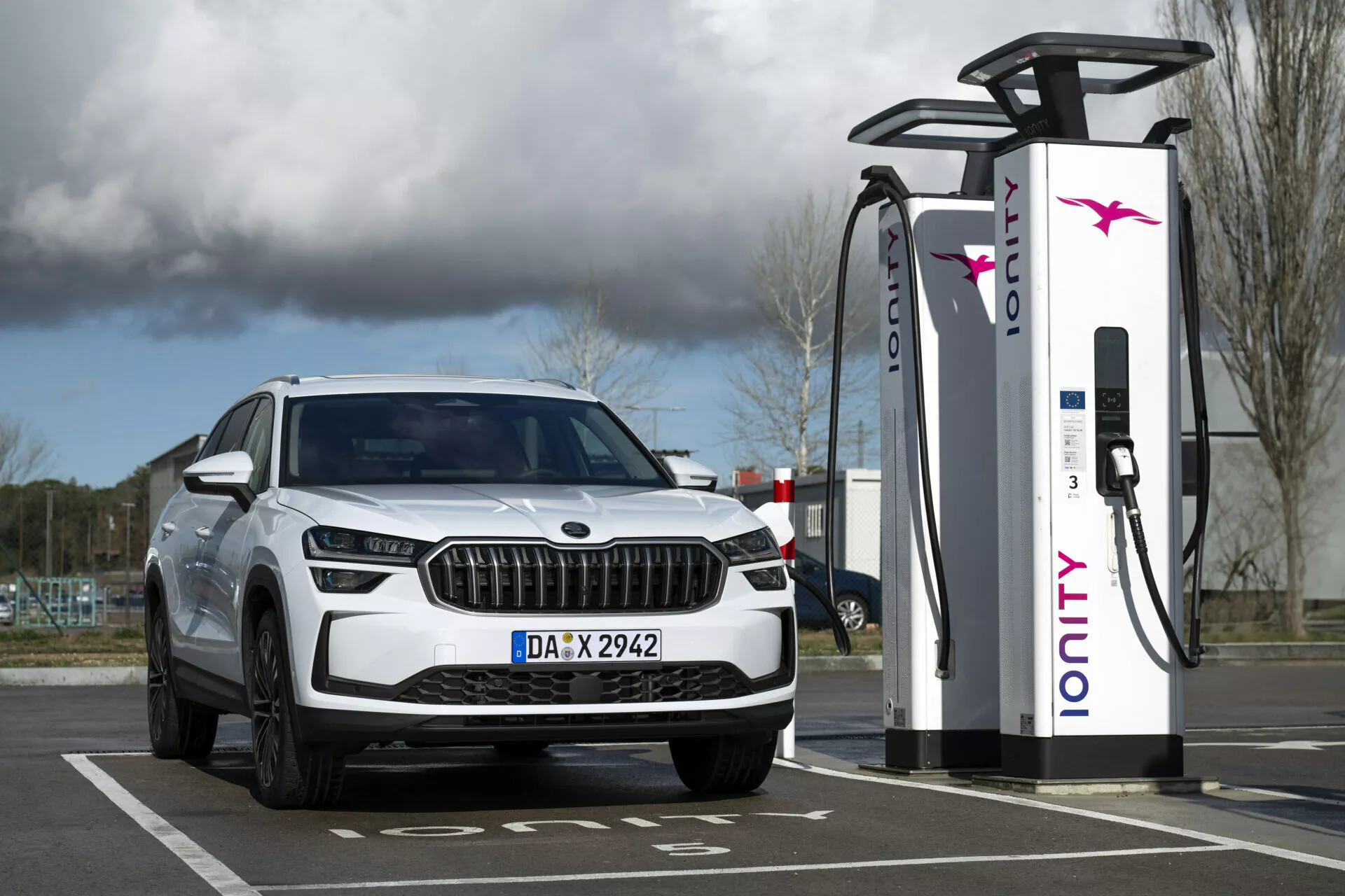 Biela SUV Škoda Kodiaq 2024 zaparkovaná pri nabíjacej stanici, zobrazenie prednej časti vozidla s LED svetlometmi a dennými svetlami, zdôrazňujúce jeho elektrické alebo hybridné schopnosti a luxusný dizajn mriežky.