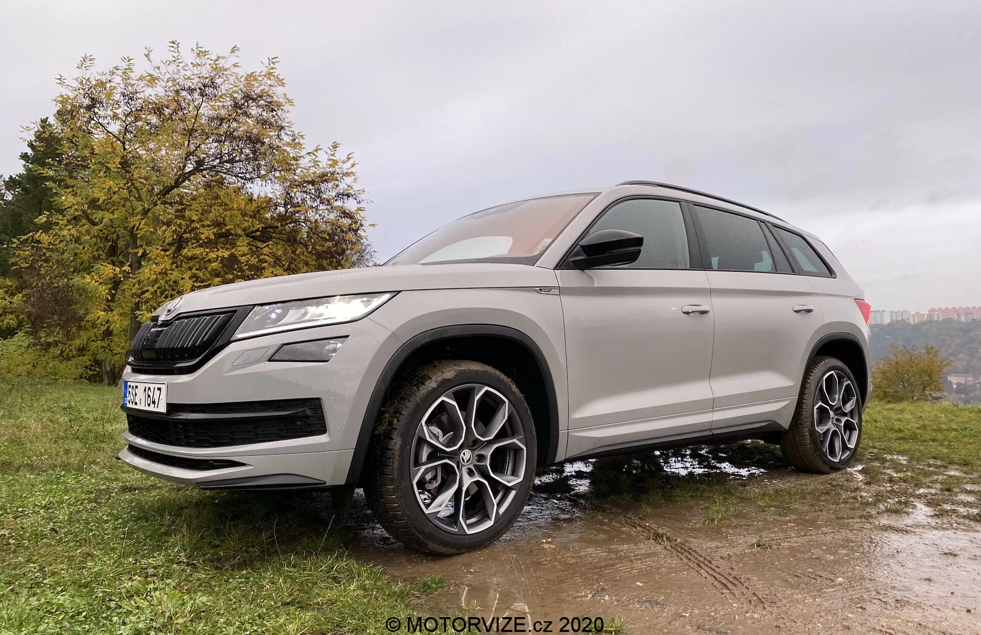 Trojštvrtinový predný pohľad na SUV Škoda Kodiaq 2017 urobený z mierne nízkeho uhla, zvýrazňujúci výšku a postoj vozidla. Vozidlo je zaparkované na nerovnom povrchu z trávy a blata, čo zdôrazňuje jeho terénne schopnosti. Škoda Kodiaq sa vyznačuje výraznou maskou chladiča a ostrými LED svetlometmi. Precízny dizajn zahŕňa veľké viacramenné zliatinové kolesá, vysokú svetlú výšku a celosezónne pneumatiky. Karoséria je natretá neutrálnou metalickou farbou s tónovanými oknami a chrómové akcenty zvýrazňujú jej prémiový vzhľad. Moderné funkcie sú viditeľné na bočných zrkadlách vo farbe karosérie, s integrovanými smerovkami a pravdepodobne aj s výstražnými systémami mŕtveho uhla.