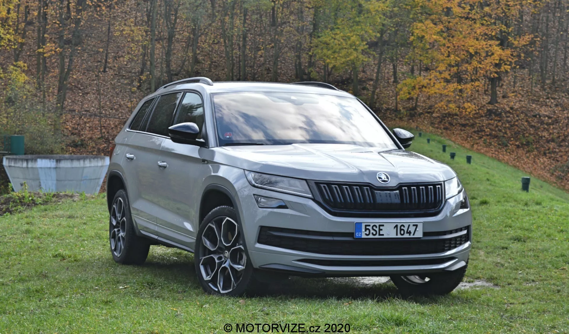 Detailný pohľad na Škoda Kodiaq 2017, strieborné SUV, umiestnené vonku na jesennej kulise stromov. Denný záber zachytáva vozidlo z predného trojštvrťového uhla, ukazujúci jeho predný a bočný profil. Viditeľné sú výrazné dizajnové prvky, vrátane prednej mriežky s logom, LED svetlometov, hmloviek, viacramenných zliatinových kolies, strešných líšt a bočných zrkadiel s integrovanými smerovkami. Auto je umiestnené na tráve blízko betónového okraja, čo naznačuje prostredie podobné parku.