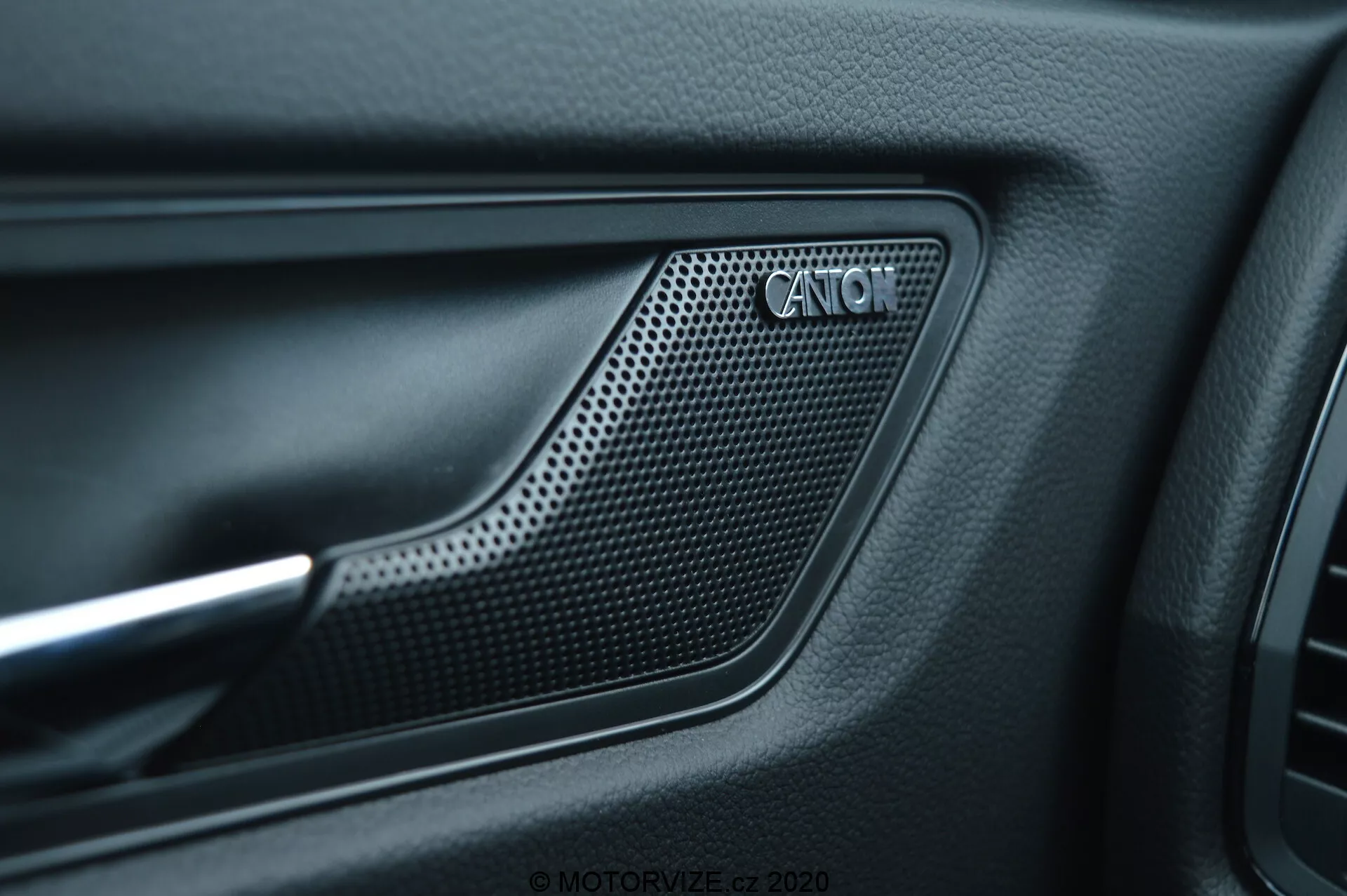 Zblízka interiérového dverového panelu auta Škoda Kodiaq 2017 s reproduktorovou mriežkou CANTON. Mriežka z perforovaného materiálu so vzorom včelieho plástu je umiestnená v zapustenej oblasti dverového panelu obkolesenej vyvýšeným obrysom. Okolo mriežky je matný čierny textúrovaný materiál a kontrastný kovový akcent, čo odráža sofistikovaný dizajn interiéru a prémiový audiosystém.