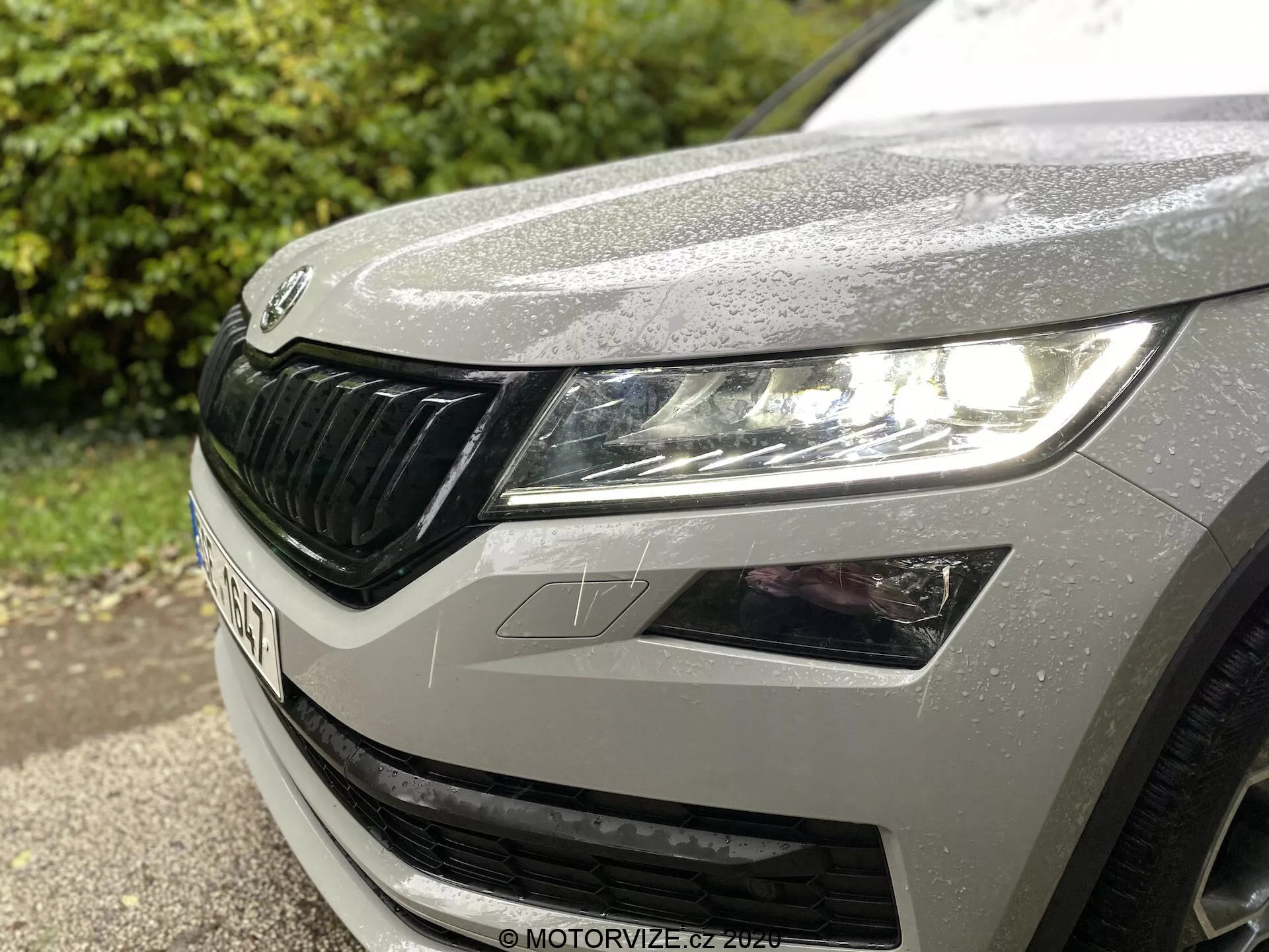 Detailný pohľad na ľavú prednú štvrtinu modelu Škoda Kodiaq 2017, zobrazujúci ľavý svetlomet s moderným LED dizajnom, časť kapoty s dažďovými kvapkami a charakteristickú mriežku Škoda s horizontálnymi prvkami.