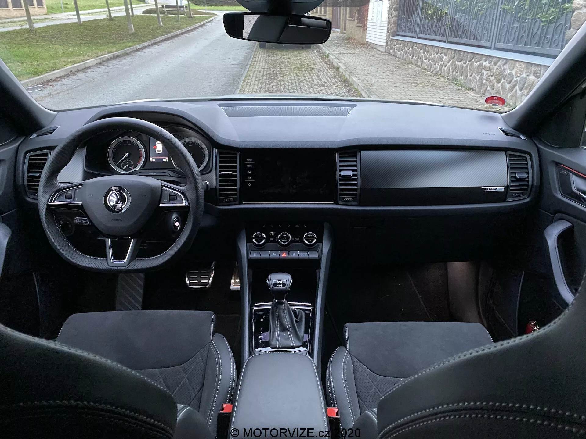 Interiér vozidla Škoda Kodiaq 2017 z pohľadu zadných sedadiel, ktorý ukazuje palubnú dosku, volant s multifunkčnými tlačidlami, digitálny displej prístrojov, široký infotainment displej, ovládanie HVAC, volič automatickej prevodovky, prémiové kožené materiály a športové sedadlá s integrovanými opierkami hlavy.