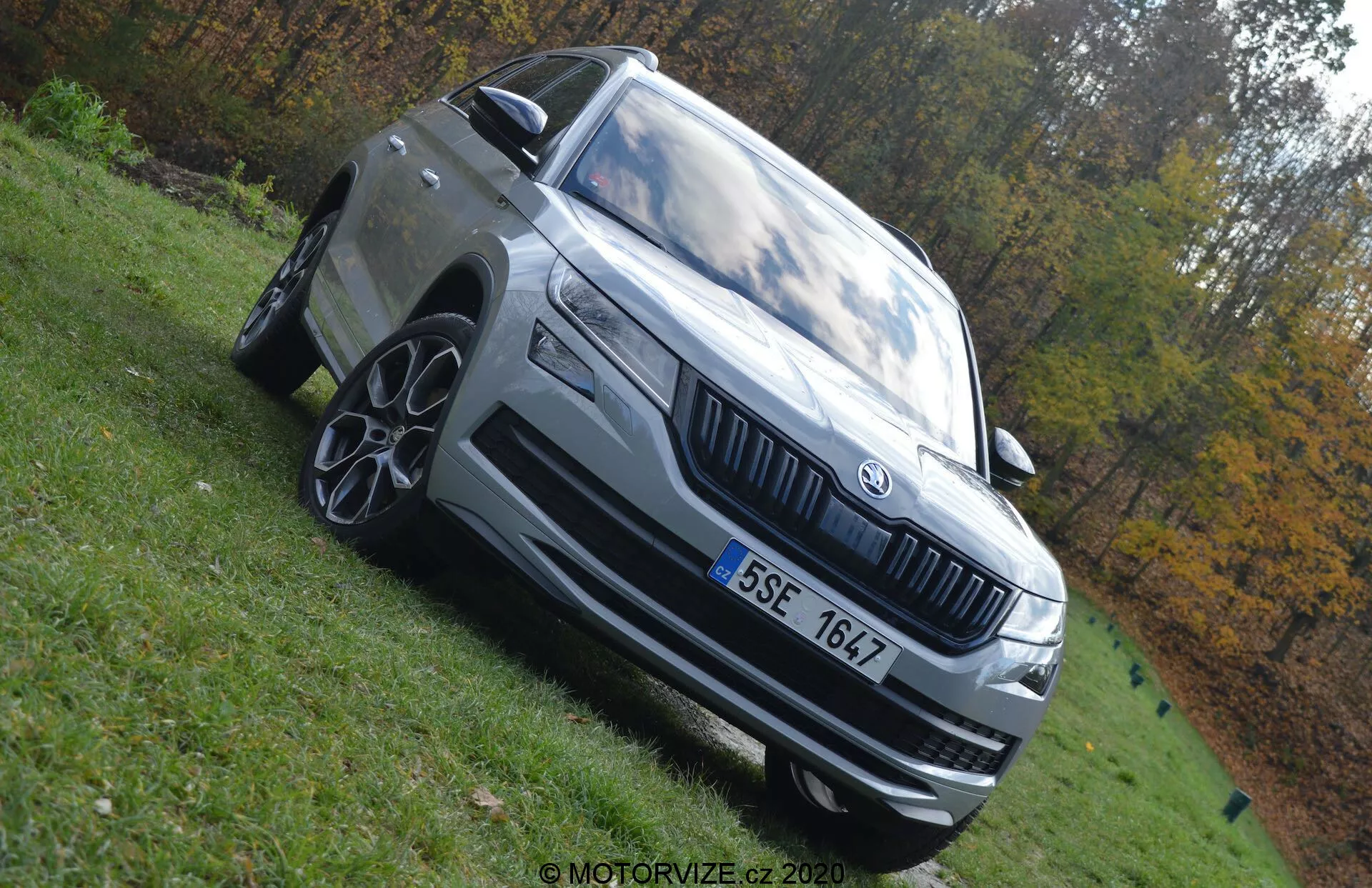 Nízkoúhlá fotografia Skoda Kodiaq 2017 zvýrazňujúca jeho dominantný postoj. Auto je zachytené so zobrazením prednej ľavej štvrtiny s viditeľnými prvkami, ako sú LED svetlomety, výrazná mriežka s logom Skoda, viaclúčové zliatinové kolesá, predný nárazník s prívodmi vzduchu, bočné zrkadlo s integrovaným smerovkami a charakteristické línie karosérie. Vozidlo je svetlo šedé alebo strieborné a je zaparkované na trávnatej ploche za denného svetla. Rozmazané jesenné pozadie zdôrazňuje prítomnosť vozidla.