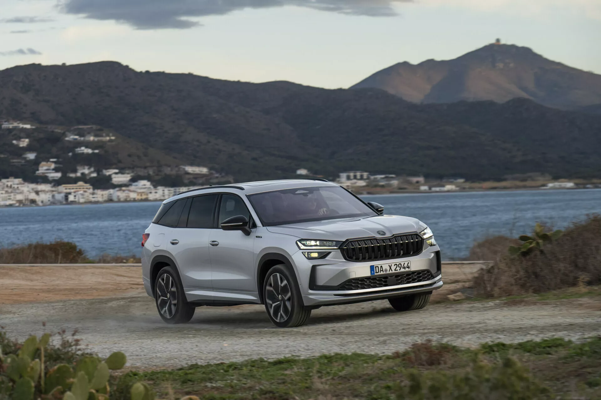 SUV Skoda Kodiaq 2024 zaparkované na nezpevnenom povrchu, s výhľadom na scénické kopce a vodnú plochu, zdôrazňujúce moderné LED svetlomety vozidla, prednú mriežku s potenciálnou integráciou ADAS a súčasný dizajn kol pod mäkkým, zamračeným denným svetlom.
