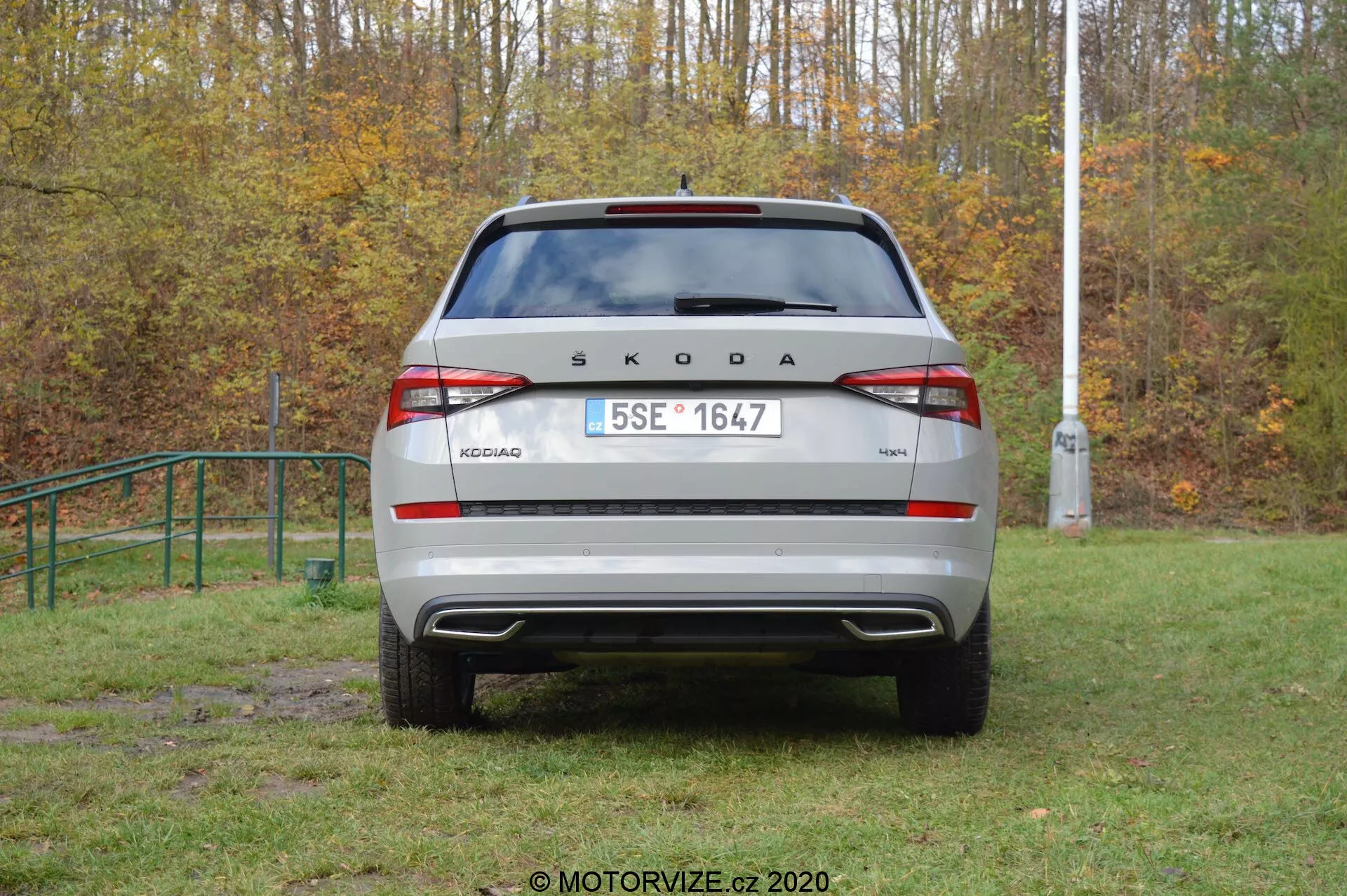 Zadný pohľad na vozidlo Škoda Kodiaq 2017 v vonkajšom prostredí. Značka 'ŠKODA' a model 'KODIAQ' sú výrazne zobrazené na veku kufra. Európska ŠPZ, moderné zadné svetlá s integrovanými hmlovými a cúvacími svetlami, viditeľný zadný nárazník s parkovacími senzormi a zadné okno so stieračom sú jasne viditeľné. Reflexné prvky na spodnej časti nárazníka zvyšujú viditeľnosť. Auto je zaparkované na trávnatej, mierne blatovej oblasti, čo naznačuje terénne schopnosti.