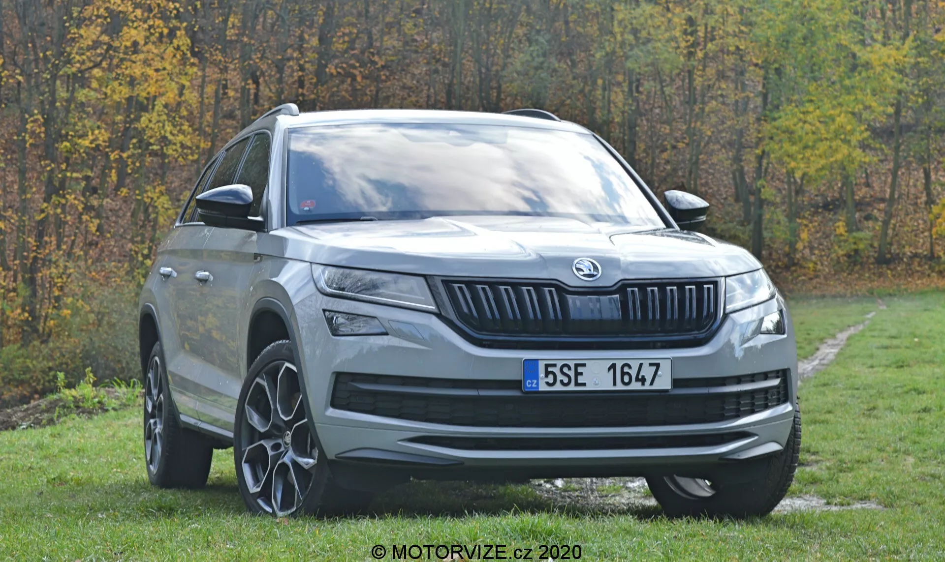 Trojštvrtinový predný pohľad na moderné SUV Skoda Kodiaq 2017 zaparkované na tráve s lesom v pozadí. Viditeľné funkcie zahŕňajú výraznú prednú masku s vertikálnymi lamelami a centrálnym logom značky, elegantné horizontálne orientované svetlomety s LED dennými svetlami, vysokú svetlú výšku, veľké viaclučové zliatinové kolesá, výrazné oblúky kolies, strieborný ochranný kryt, výrazné línie kapoty, teleskopické spätné zrkadlá s integrovanými smerovkami, tmavené bočné okná, mierne zníženú líniu strechy smerom dozadu so strešnými nosičmi.