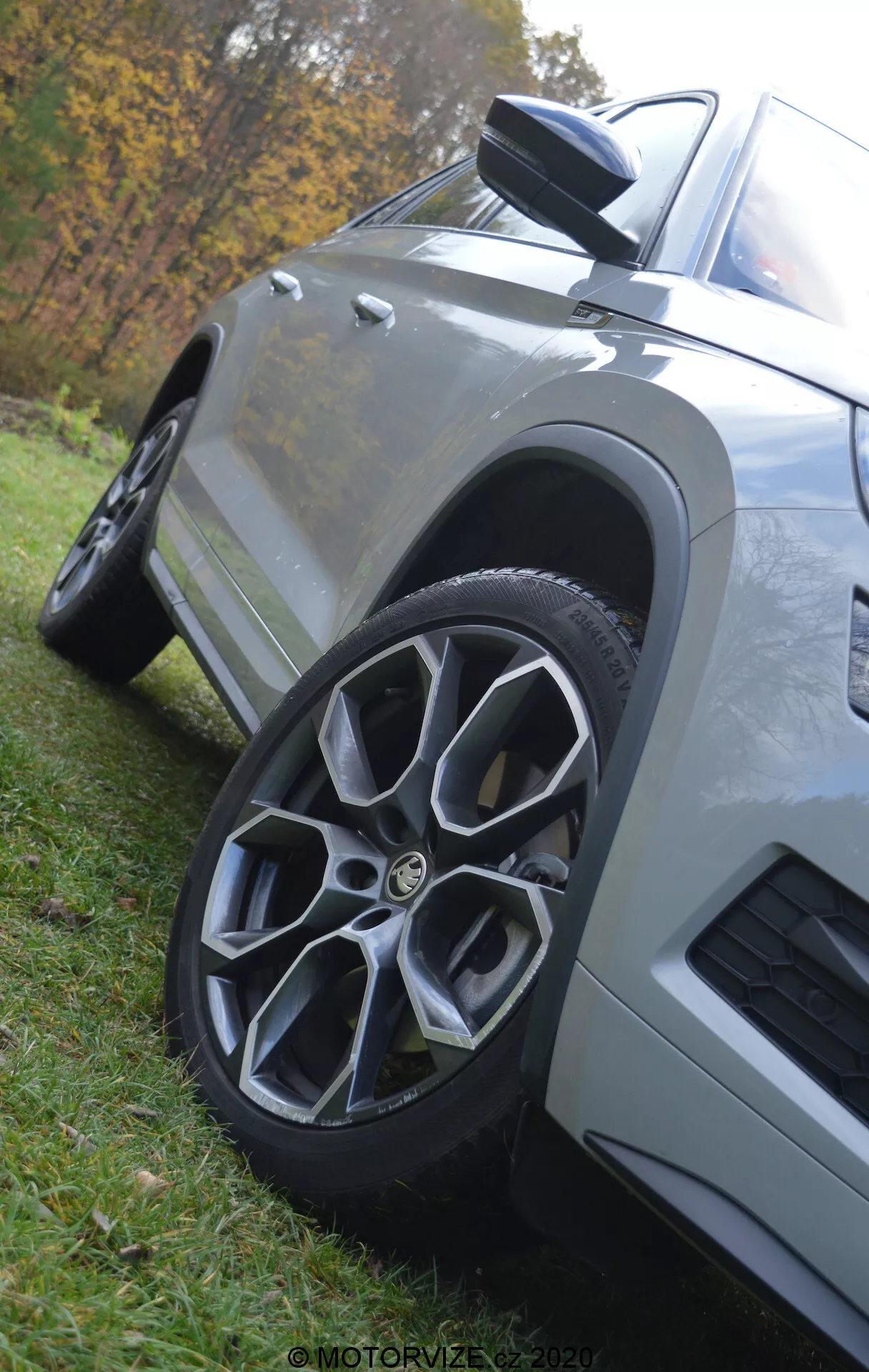 Detailný záber z nízkeho uhla na Škoda Kodiaq 2017, zvýrazňujúci predné koleso a blatník. Auto zaparkované na tráve má viacramenné zliatinové koleso, aerodynamické bočné zrkadlo s integrovaným indikátorom, hranaté karosárske panely s metalickým lakom a prieduch na prednom blatníku, všetko na rozmazanom prírodnom pozadí.