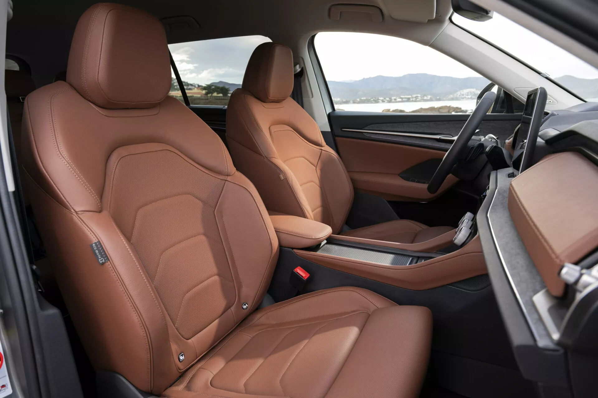 Interiér Škoda Kodiaq 2024 s luxusnými prednými sedadlami potiahnutými hnedou kožou s detailným prešívaním, elektrickým nastavením sedadla a pohľadom na prvky vysokej kvality ako sú športovo inšpirované bočné opierky, čierno-hnedá dvojfarebná palubná doska a minimalistická stredová konzola s automatickou radicou pákou, z pohľadu otvorených dverí na strane spolujazdca