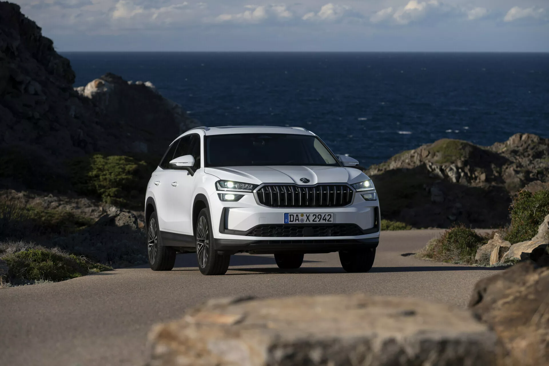 Biely SUV Skoda Kodiaq 2024 zaparkovaný na pobrežnej ceste s oceánom na pozadí, s výraznou prednou mriežkou s vertikálnymi lištami podobnými chrómu, dennými svetlami LED, diskami z ľahkej zliatiny s viacerými lúčmi a detailným pohľadom z predného trojštvrte úhla