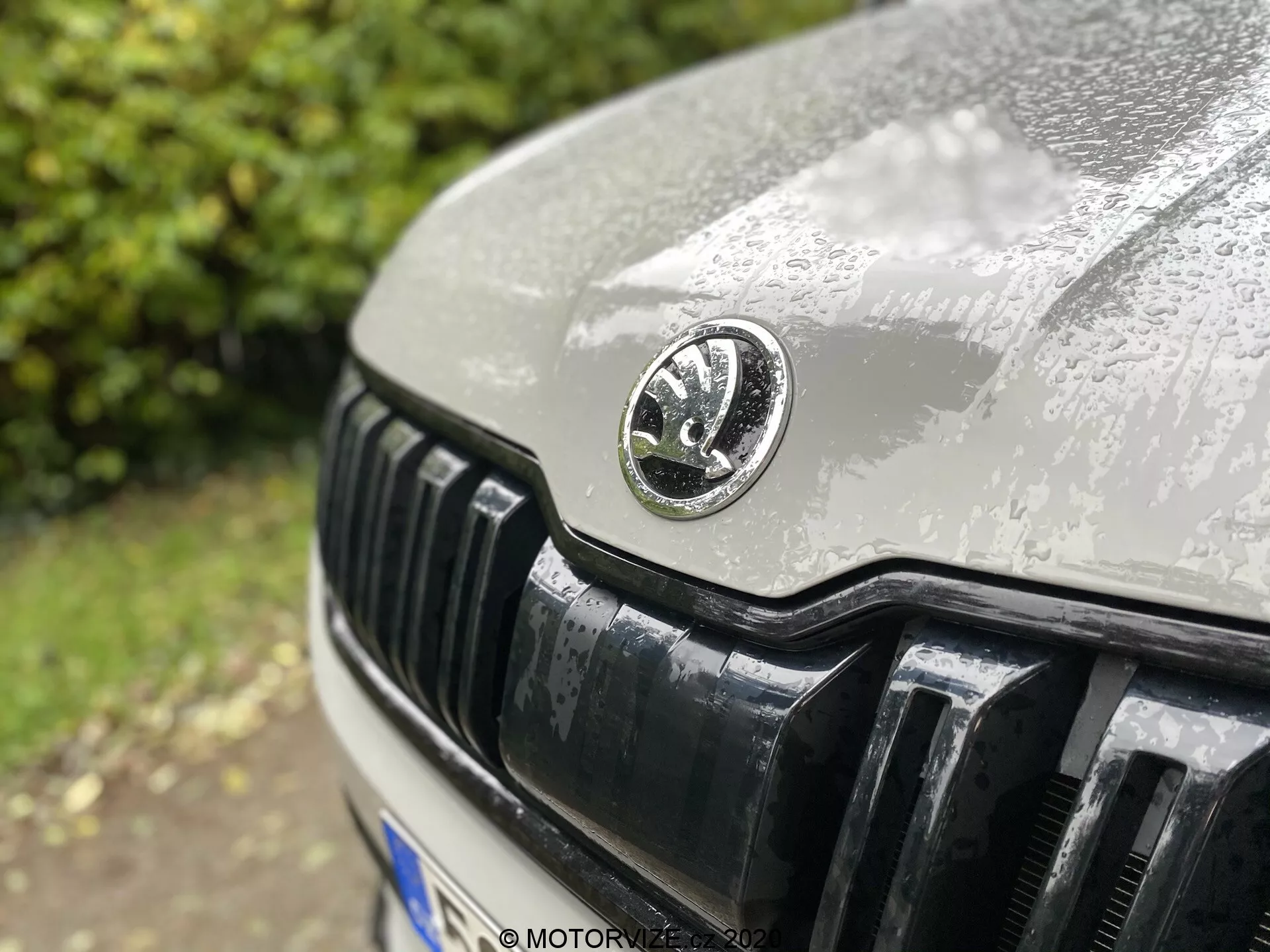 Detailný záber prednej časti vozidla Skoda Kodiaq 2017, ktorý sa zameriava najmä na mriežku chladiča a emblém výrobcu. Kruhový emblém je umiestnený v strede, zobrazujúc jemné detaily s kvapkami vody, ktoré naznačujú nedávny dážď. Mierne mimo stredový uhol snímky zachytáva kapotu a lesklé čierne vertikálne lamely mriežky v ostrom zábere s plytkou hĺbkou ostrosti.