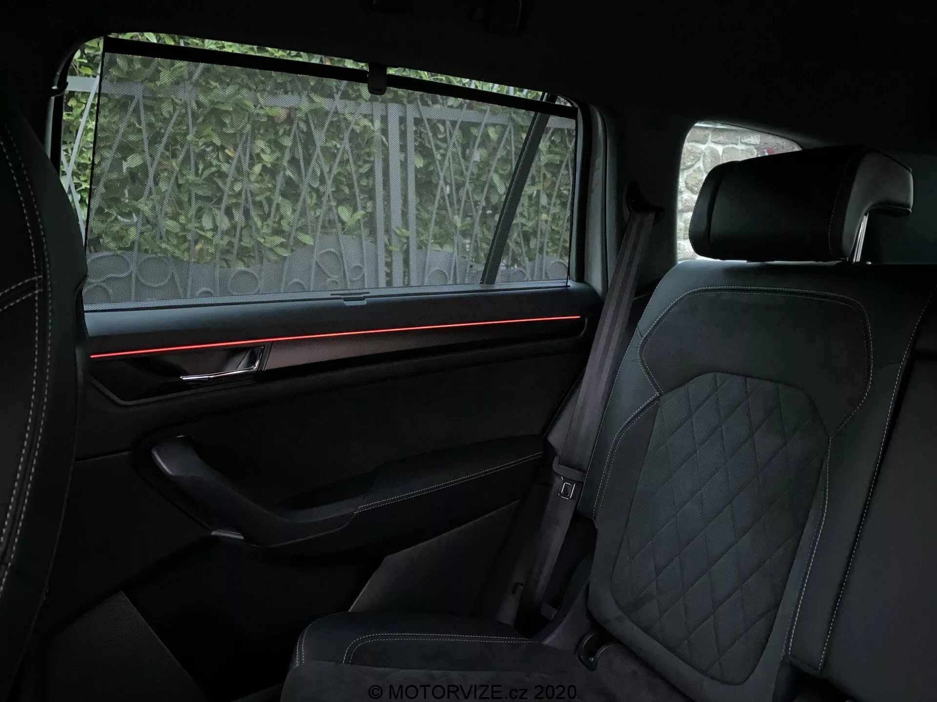 Interiérový pohľad na Skoda Kodiaq 2017 zameraný na zadný priestor pre cestujúcich, zobrazujúci luxusné prvky ako prešívané čalúnenie z kože na predných aj zadných sedadlách, červený ambientný osvetlený pásik na dobre vybavenom polstrovanom paneli dverí s prémiovým obkladom a čiastočne priehľadná slnečná clona na okne. Dôraz je kladený na pohodlie a prvotriedne dizajnové prvky.
