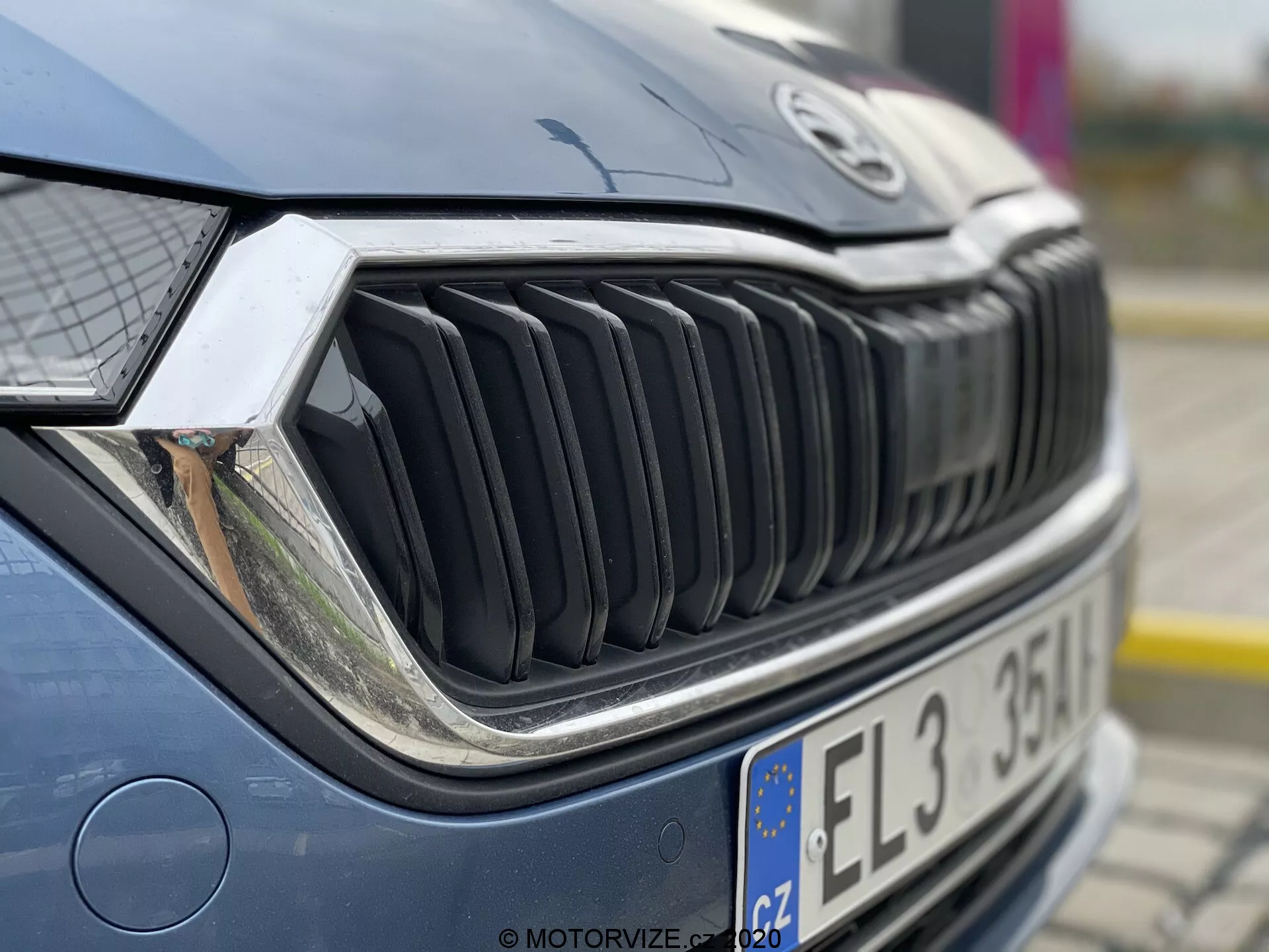 Detailný záber na prednú časť auta so zameraním na mriežku chladiča a svetlomet. Fotografia je urobená z predného pravého uhla, mierne nahor a ukazuje detailný pohľad na pravú stranu mriežky, pravý svetlomet a časť kapoty. Vertikálne lamely mriežky sú výrazné, naznačujúce odvážny dizajn. Složitý svetlomet naznačuje pokročilú svetelnú technológiu, ako sú LED alebo adaptívne systémy. Kapota je mierne rozmazaná, zdôrazňujúc textúru mriežky a svetlometu. Auto je dobre udržiavané, s odrazmi naznačujúcimi vonkajšie prostredie v zamračený deň alebo v tieni.