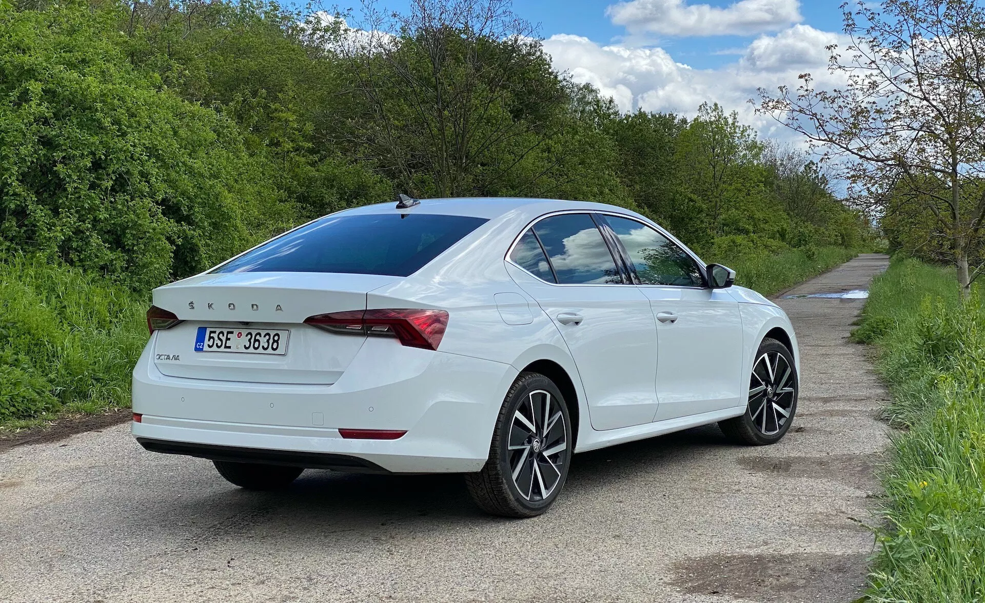 Zadný trojštvrťový pohľad na biely sedan Skoda Octavia 2020 zaparkovaný na pravej strane úzkej cesty, zobrazujúci stranu spolujazdca z úrovne zeme. Auto zaberá väčšinu rámu, zdôrazňuje dizajnové línie, zadné svetlá obklopujúce kufor, jemne integrovaný spojler, zadné označenie, viacramenné zliatinové kolesá, mierne zatmavené zadné okno a bočné zrkadlá s integrovanými smerovkami.