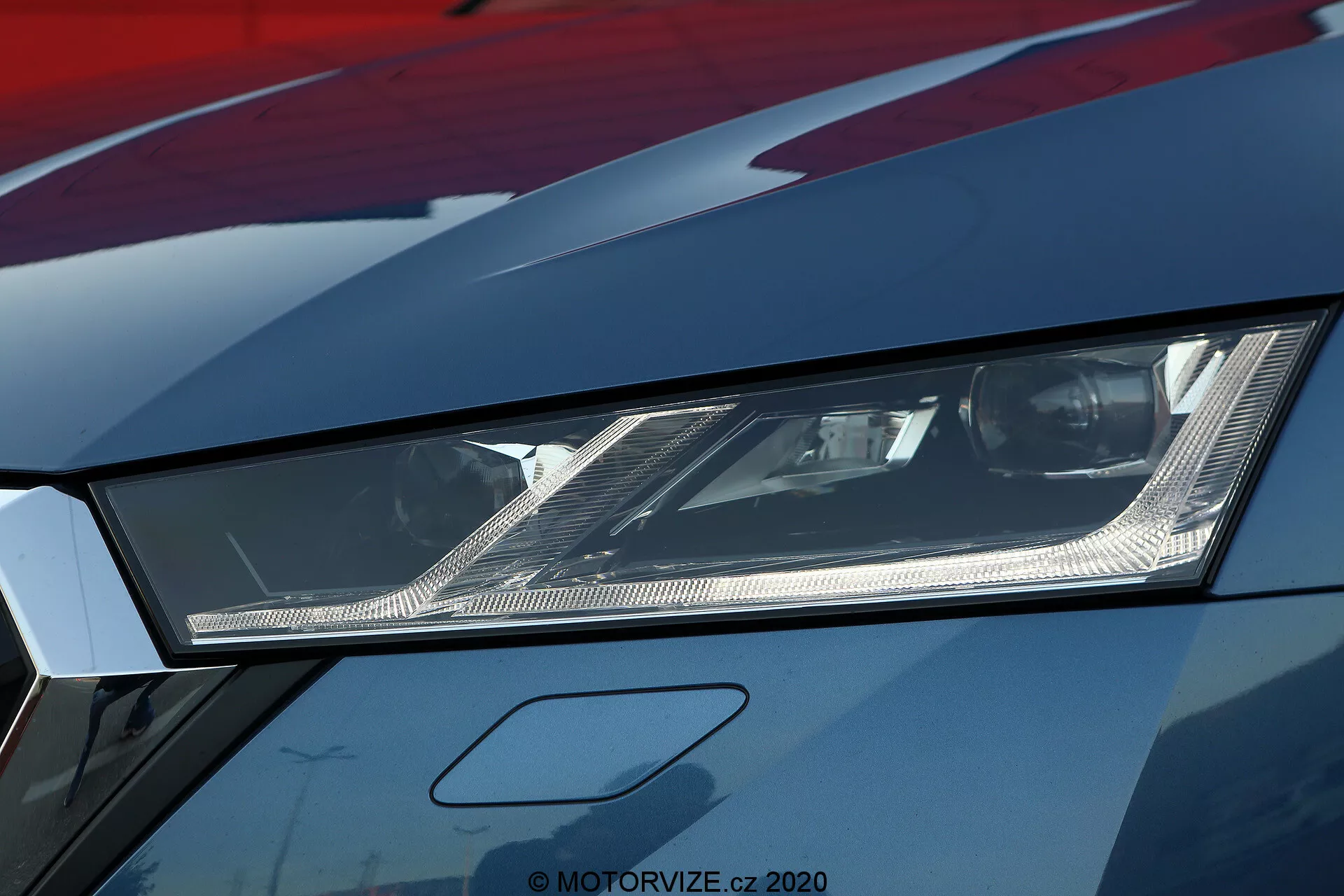 Detailný záber na ľavý svetlomet modelu Škoda Octavia 2020 limuzína, zvýrazňujúci jeho hranatý dizajn a modernú LED alebo laserovú osvetľovaciu technológiu. Fotografia, urobená z nízkeho uhla, ukazuje zložité vnútorné reflektory a jemné detaily vo svetlomete pokrytom čírou šošovkou. Vysoké rozlíšenie odhaľuje aj časť kapoty a blatníka s kovovou povrchovou úpravou, čo naznačuje luxusné alebo dobre udržiavané vozidlo. Tento obrázok zdôrazňuje pokročilé automobilové osvetlenie a agresívny štýl spojený so športovými autami.