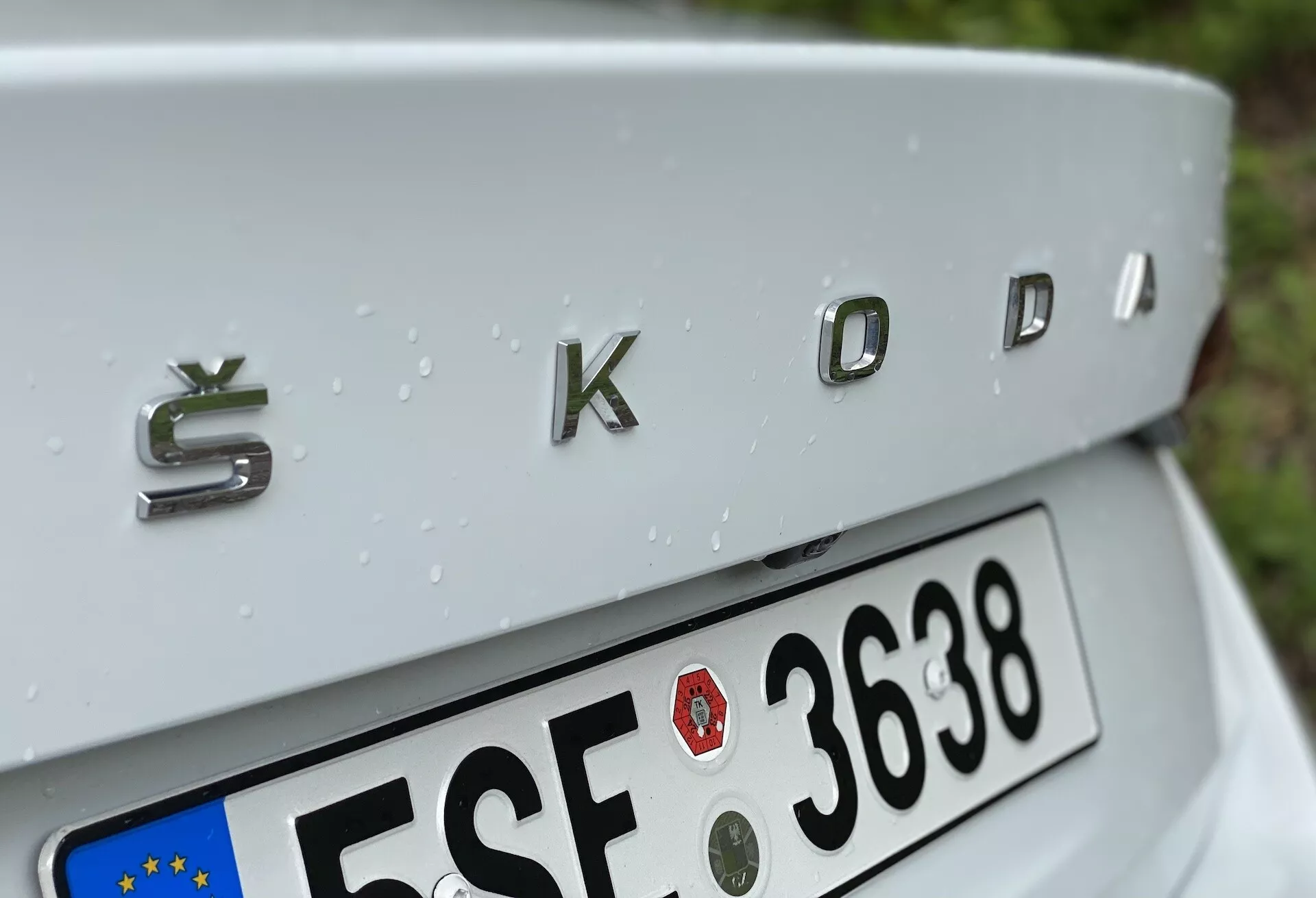 Detailný záber zadnej časti Škoda Octavia 2020 (octavia-limousine) so zameraním na chrómovaný znak 'ŠKODA' s trojrozmerným kovovým povrchom. Uhol záberu je mierne zhora nadol, ukazuje jemne sa zvažujúce veko kufra, čiastočne viditeľnú európsku tabuľku s evidenčným číslom pod ním a kvapky dažďa na striebornom povrchu auta, čo naznačuje nedávny dážď.