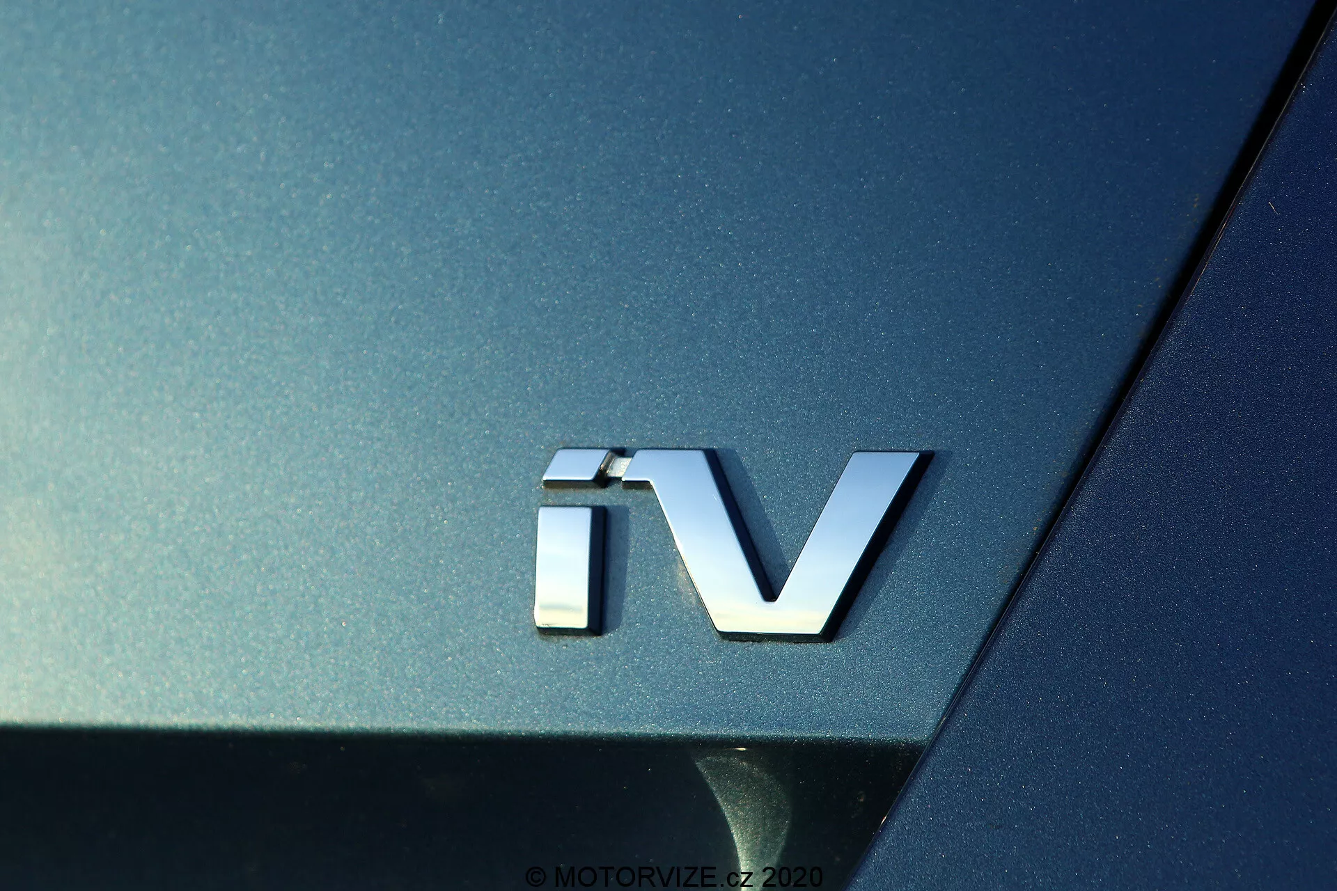 Detailný záber na znak 'iV' modelu Škoda Octavia 2020, poukazujúci na elektrifikovaný pohonný systém, so strieborným 3D emblémom na tmavom kovovom povrchu, zvýrazňujúci krivky a odrazivosť zadnej časti vozidla.