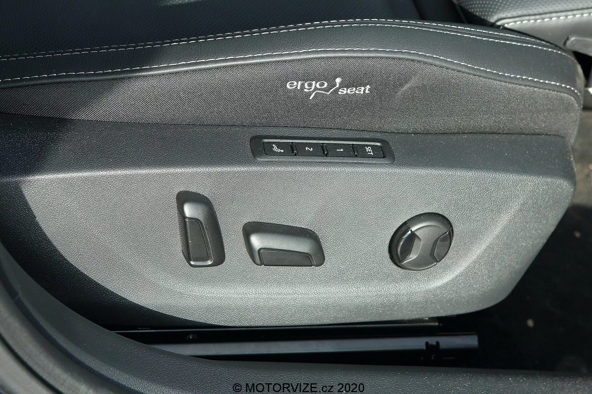 Záber zblízka predvádzajúci ovládanie sedadla ergo systému vo vozidle Škoda Octavia Limousine z roku 2020. Fotografia zdôrazňuje niekoľko tlačidiel a otočný ovládač na nastavenie polohy, výšky, sklonu a bedrovej opierky sedadla vodiča alebo spolujazdca. Nad ovládaním je viditeľne vyšitý nápis 'ergo seat', čo zdôrazňuje ergonomický dizajn. Sedadlo je z tmavosivej tkaniny, naznačujúcej odolný a pohodlný materiál vhodný pre vozidlá strednej a vyššej triedy.