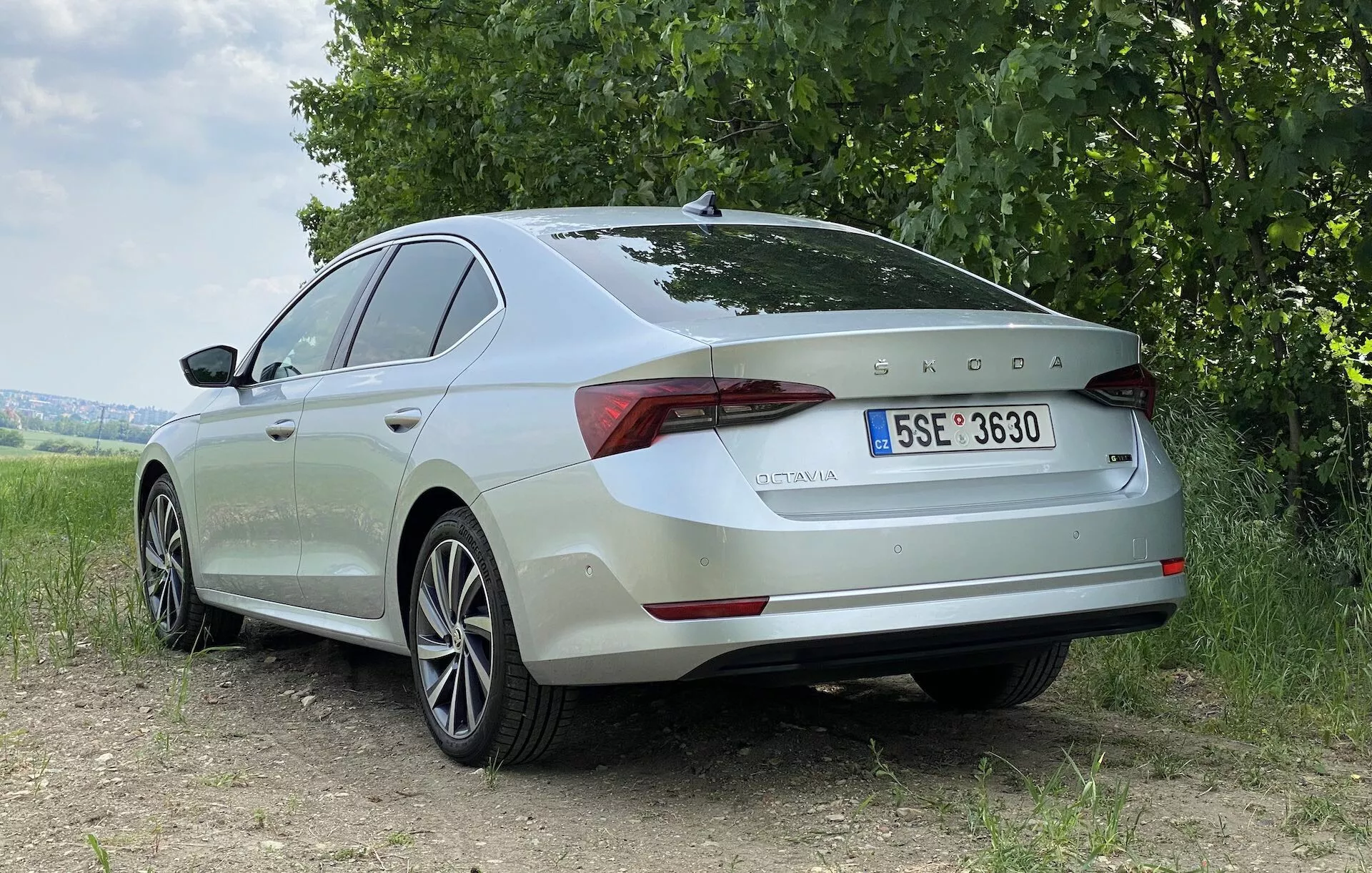 Pohľad na zadných tri štvrtiny svetlo zfarbeného sedanu Škoda Octavia 2020 na špinavej ploche s trávou a stromami v pozadí. Fotografia zdôrazňuje zadný a bočný profil vozidla, ukazuje hladké línie, tvarované povrchy, hliníkové kolesá a elegantné LED zadné svetlá. Na veku kufra sú jasne viditeľné znaky Škoda a modelové označenie 'OCTAVIA', spolu s európskou registračnou značkou. Prirodzené, mäkké svetlo zvýrazňuje dizajnové prvky auta bez ostrých tieňov, čo naznačuje zatiahnutú oblohu alebo rozptýlené slnečné svetlo.