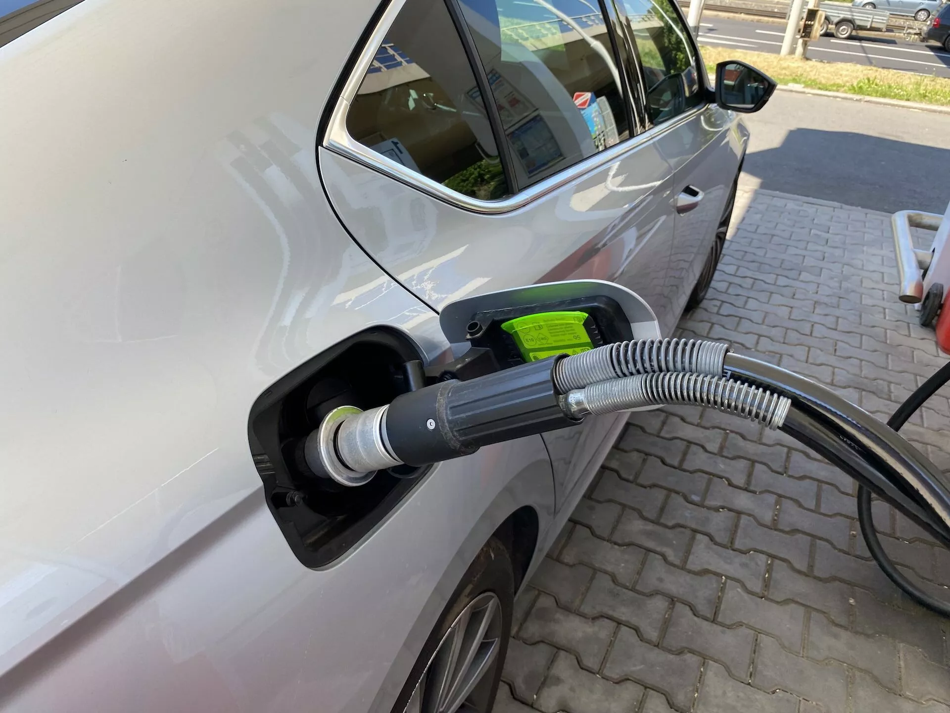 Detailný pohľad na ľavú stranu Škody Octavia 2020 pripojenú k elektrickej nabíjacej stanici, zvýrazňujúc oblasť nabíjacieho portu so zameraním na nabíjací kábel a port. Obrázok je nasnímaný z mierneho uhla od prednej časti smerom k zadnej, zobrazujúc predné dvere vodiča a časť zadných dverí, čo naznačuje, že ide o vozidlo viacerými dverami. Vlastnosti zahŕňajú moderné bočné zrkadlo, čiastočne viditeľný zliatinový disk, svetlú farbu auta s lesklým povrchom, otvorený nabíjací port s hrubým, vlnitým ochranným krytom na kábli a zelenú rukoväť zástrčky symbolizujúcu ekologickosť. Auto je zaparkované na dláždenej ploche so vzorom tehál, čo naznačuje vyhradenú nabíjaciu stanicu, bez iných vozidiel v okolí, čo zdôrazňuje proces nabíjania.
