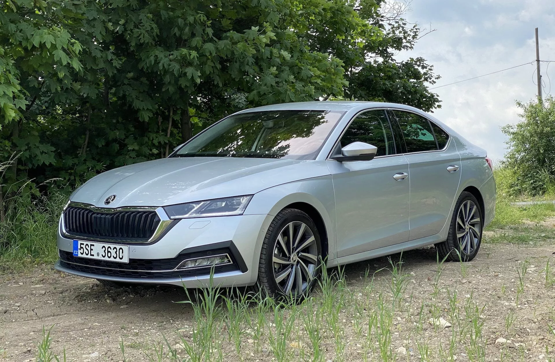 Skoda Octavia 2020 zobrazená z nízkeho čelného trojštvrtinového uhla, zobrazujúca detailne prednú ľavú stranu vrátane LED svetlometu, výraznej mriežky, predného nárazníka so senzormi, vyrezávaných línií kapoty, viacramenných zliatinových kolies, integrovaného smerovky v bočnom spätnom zrkadle a mierne tónovaných okien, umiestnená na nespevnenom povrchu so zeleným lístím a drôteným plotom vo vidieckom alebo polo-vidieckom prostredí.