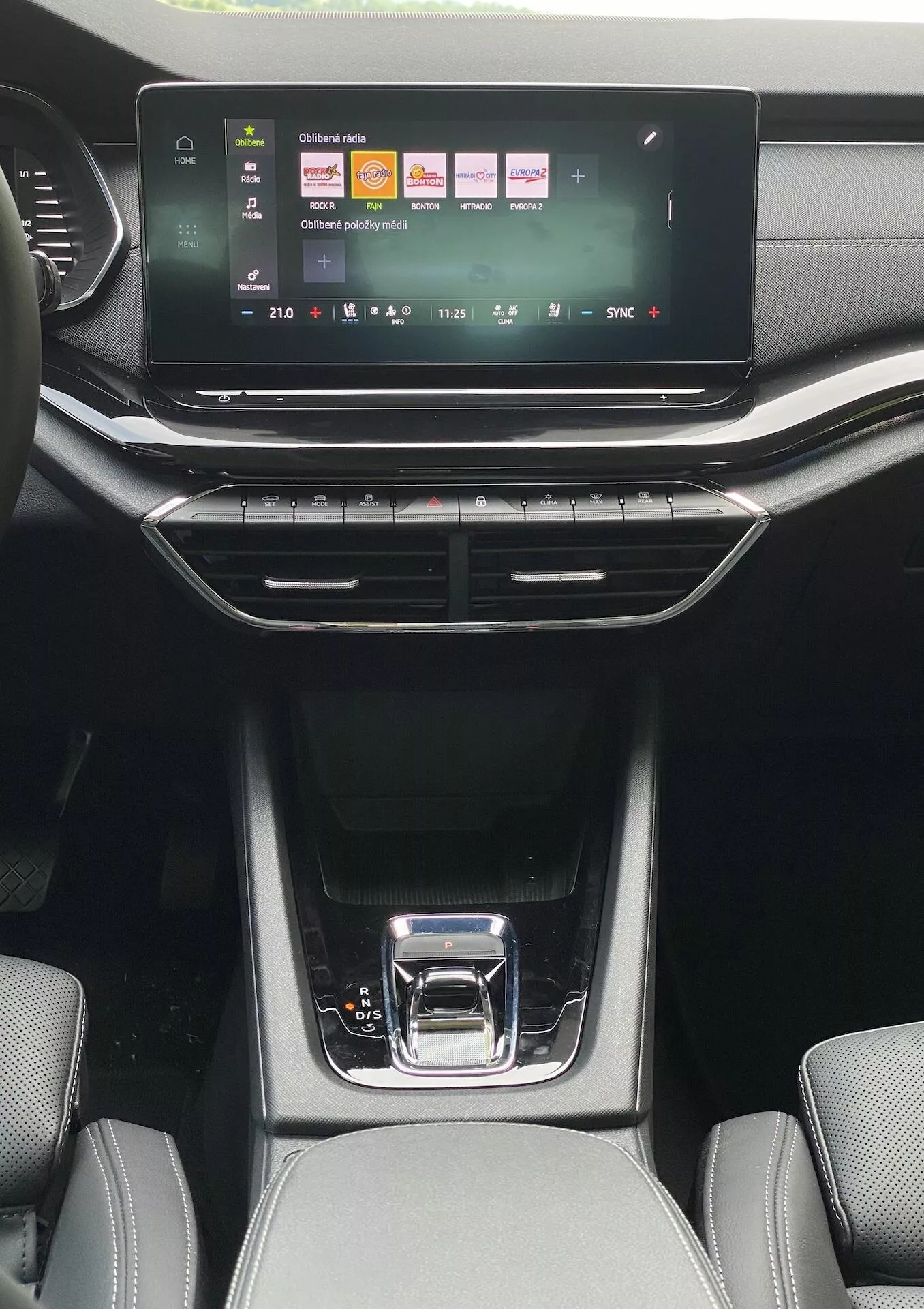 Pohľad na interiér z priestoru zadných sedadiel vozidla Skoda Octavia limousine z roku 2020, ktorý zobrazuje dotykový infotainment systém s farebnými ikonami na média, telefón a nastavenia vozidla. Pod ním sa nachádzajú tlačidlá dvojzónovej klimatizácie s digitálnym zobrazením teploty, ktoré sú obkolesené vertikálnymi a horizontálnymi výdychmi vzduchu. Elegantná stredová konzola obsahuje lesklú čiernu radiacu páku so striebornou rukoväťou, úložné priestory s gumovými rohožami a viditeľným textúrovaným čalúnením predných sedadiel pod prirodzeným denným svetlom.