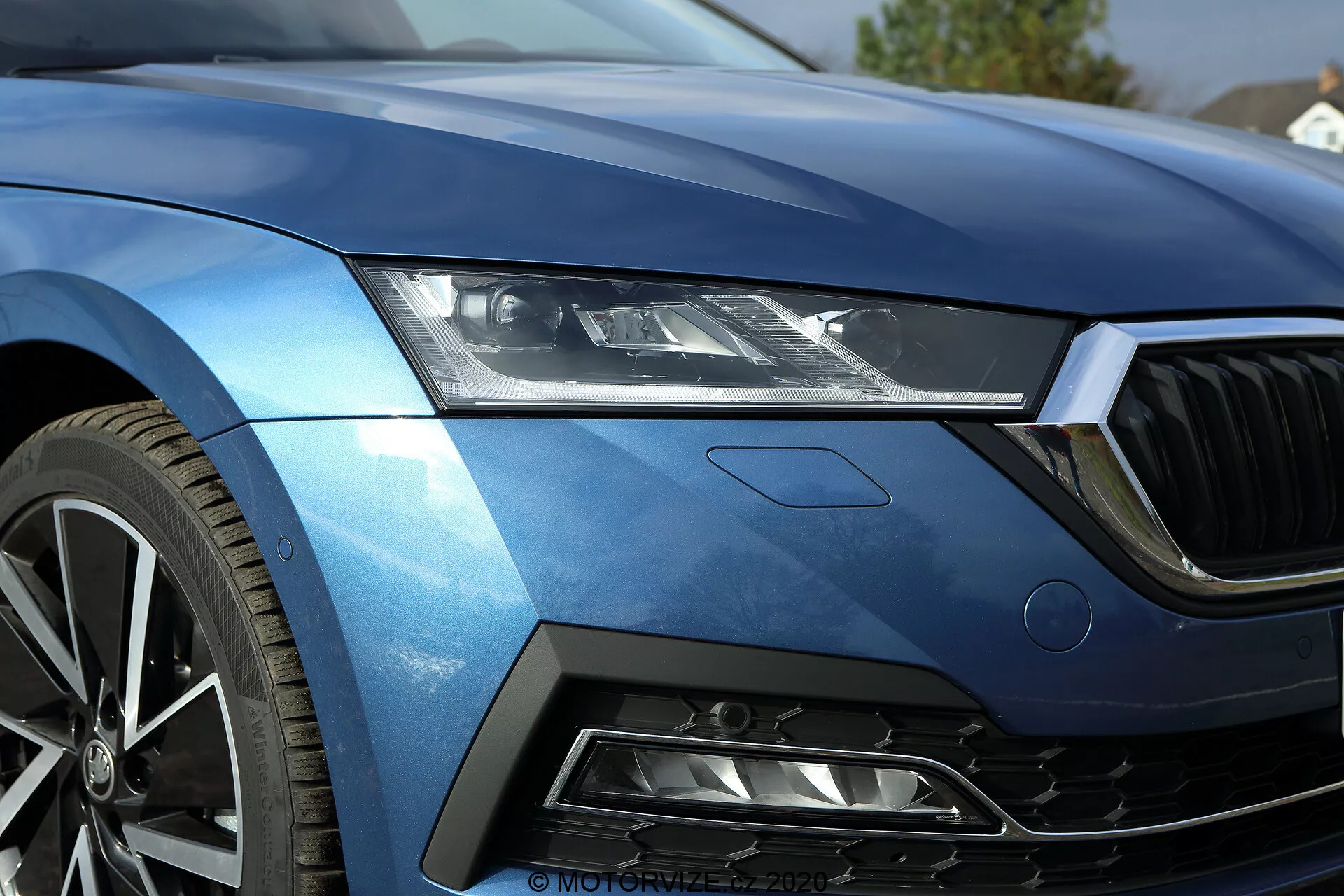 Zblízka predného ľavého štvrťroku limuzíny Škoda Octavia 2020, zobrazujúce elegantné LED reflektory, časť kapoty, ľavý okraj prednej mriežky, aerodynamický predný ľavý blatník a segment nárazníka s sací otvor s medovým očkom. Kovovo modrý lak a ostré línie zvýrazňujú luxus a športovosť. Viacramenné zliatinové koleso naznačuje špičkový výkon. Dizajn obsahuje jemný odznak, naznačujúci konkrétnu úroveň výbavy alebo technologický balík. Tento obrázok krásne zachytáva pokročilú technológiu osvetlenia vozidla, povrchovú úpravu a výkonovo orientované dizajnové prvky.