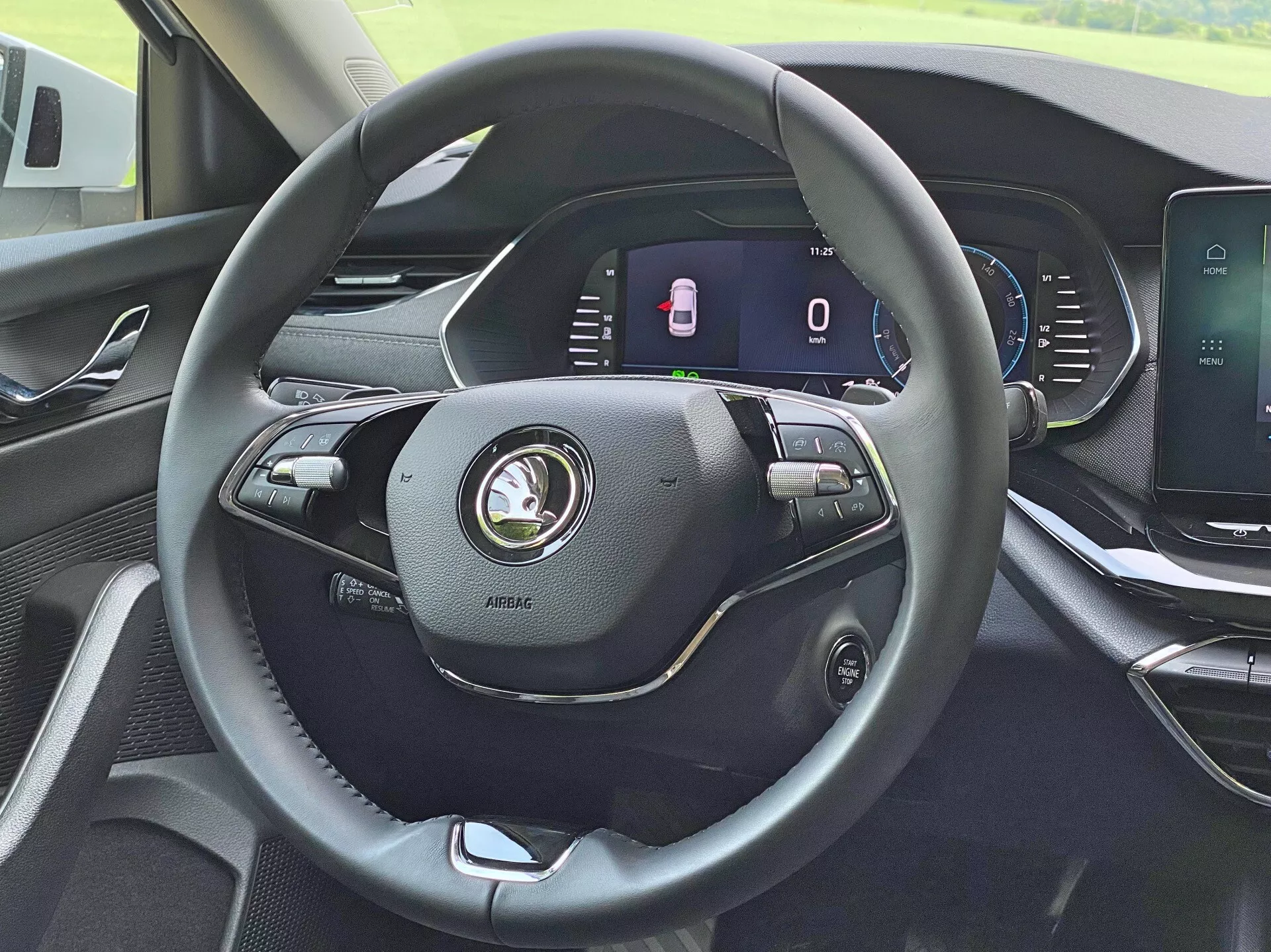 Interiér vozidla Skoda Octavia Limousine 2020 z pohľadu vodiča, zdôrazňujúci moderný trojramenný volant potiahnutý kožou s rôznymi ovládacími prvkami pre hlasitosť, hlasové príkazy a tempomat. Plne digitálny prístrojový panel za volantom zobrazuje stojaci rýchlomer a ukazovateľ otvorených dverí. Stredová konzola obsahuje dotykový infotainment displej pre multimédiá, navigáciu a nastavenia vozidla. Palubná doska ukazuje moderný dizajn s vysoko kvalitnými materiálmi.