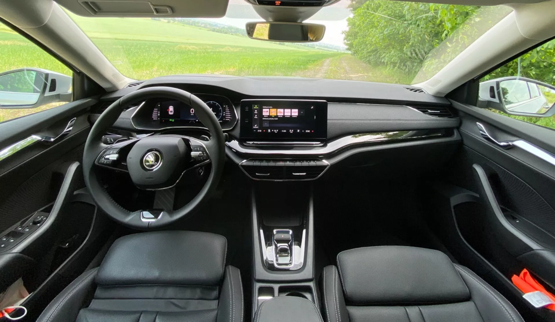 Pohľad na interiér Škoda Octavia 2020 z zadného sedadla zobrazujúci moderný volant so splošteným spodkom a integrovanými ovládačmi, digitálnu prístrojovú dosku, veľký dotykový infotainment systém na elegantnej palubnej doske, minimum fyzických tlačidiel, bezdrôtovú nabíjaciu podložku, ergonomické čalúnené predné sedadlá s kontrastným prešitím a panoramatické strešné okno vpúšťajúce prirodzené svetlo do kabíny.