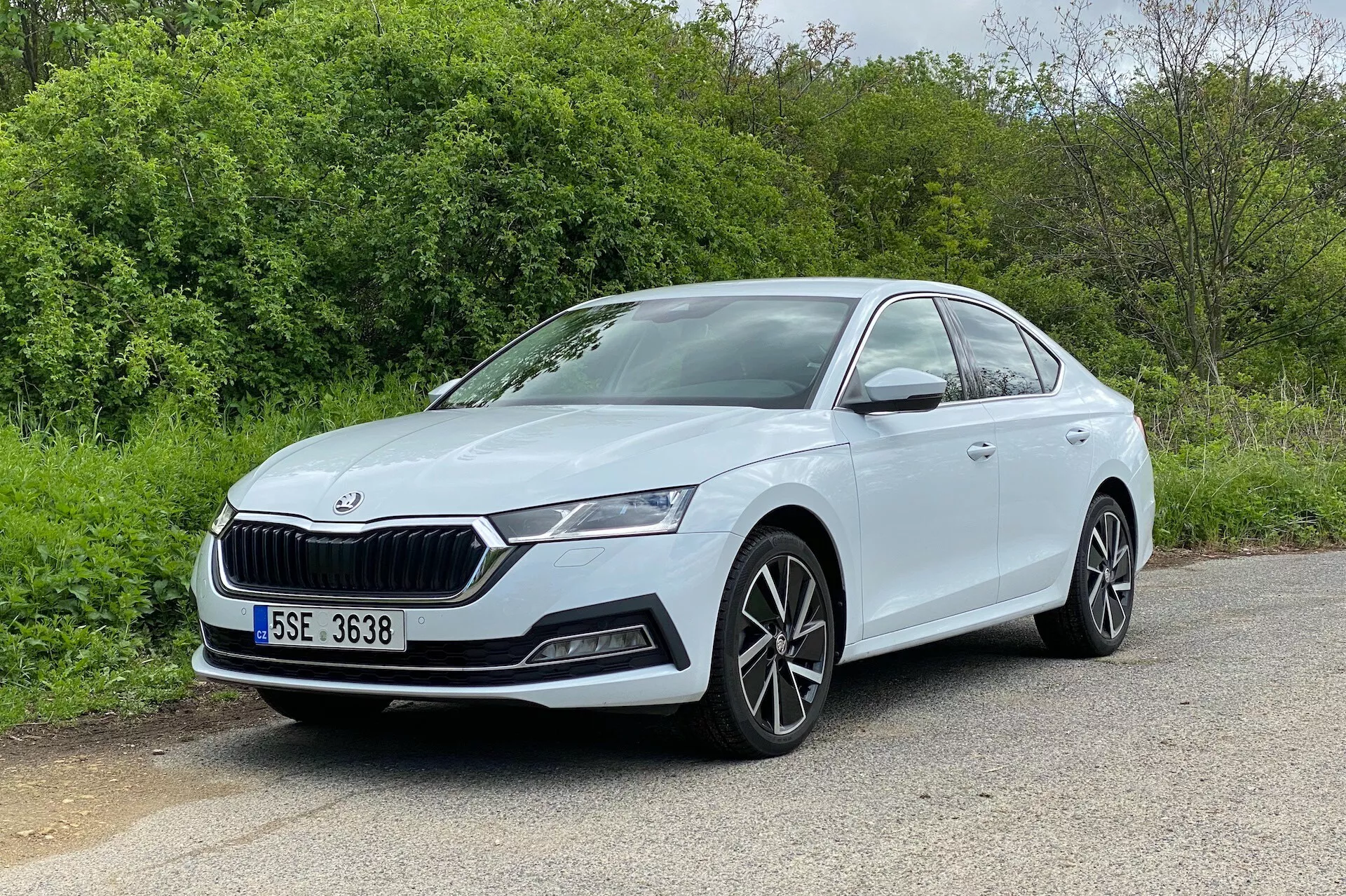 Obrázok zobrazuje sedan Škoda Octavia 2020 v uhloch predného trojštvrťového pohľadu, ktorý ukazuje prednú časť a bočný profil na strane vodiča. Auto, zaparkované na asfaltovom povrchu s prírodnými prvkami ako sú stromy alebo kríky v pozadí, má charakteristickú mriežku, elegantné LED svetlomety, hmlové svetlá a nasávacie otvory na nárazníku. Viacramenné zliatinové kolesá a hladké, aerodynamické bočné línie zvyšujú jeho moderný a športový vzhľad. Dobře osvetlený a neobmedzený výhľad zvýrazňuje farebne lakované kľučky dverí a viditeľnú charakterovú linku pozdĺž dverí, čo z neho robí vynikajúcu reprezentáciu automobilového dizajnu.